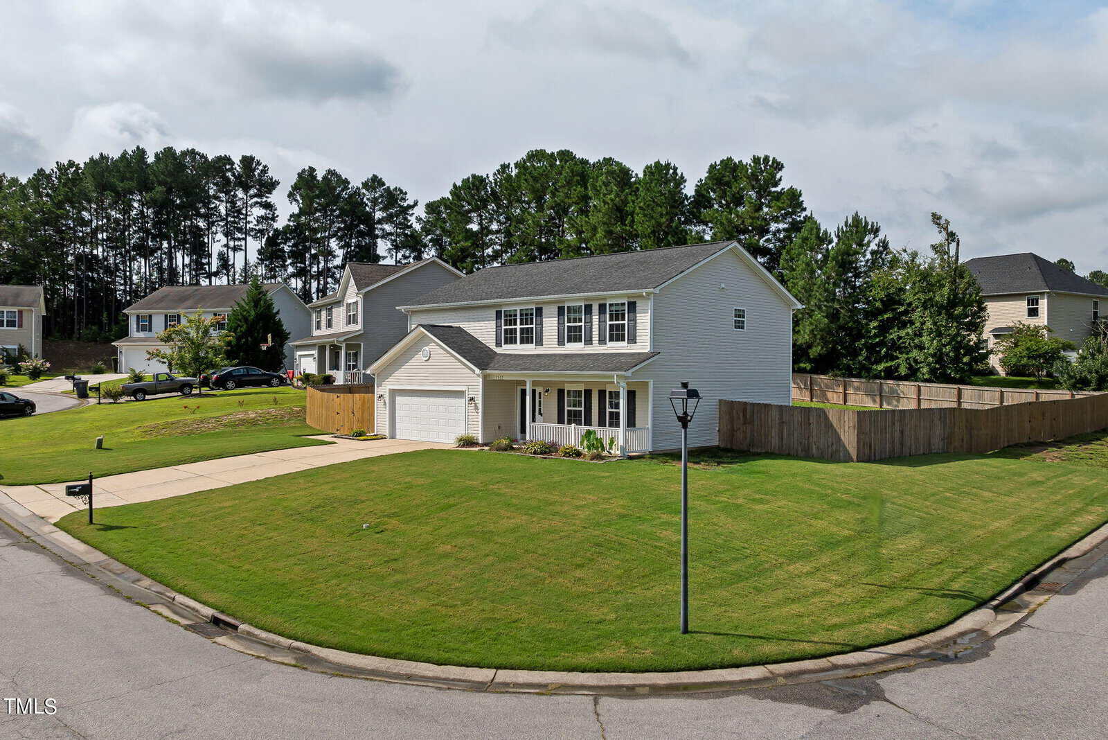 A suburban neighborhood featuring a two-story gray house with a two-car garage, white trim, and a neatly manicured lawn. The house is situated on a corner lot with a wooden fence and a streetlamp nearby, surrounded by other similar homes and tall trees.