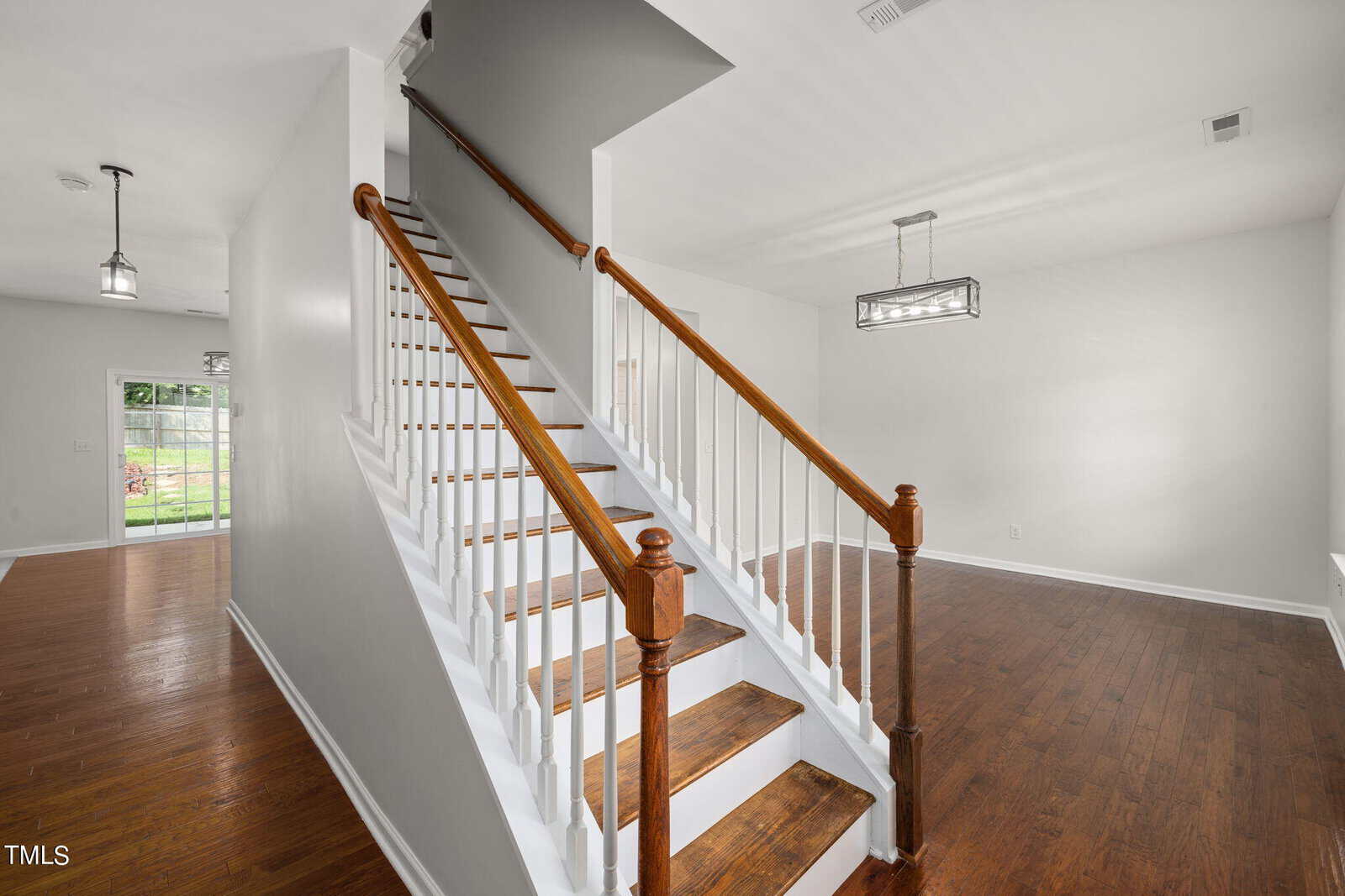 A modern interior featuring a wooden staircase with white risers and railings. It leads to the upper floor of an open living space with hardwood floors. A large window and contemporary light fixtures enhance the bright, airy ambiance.