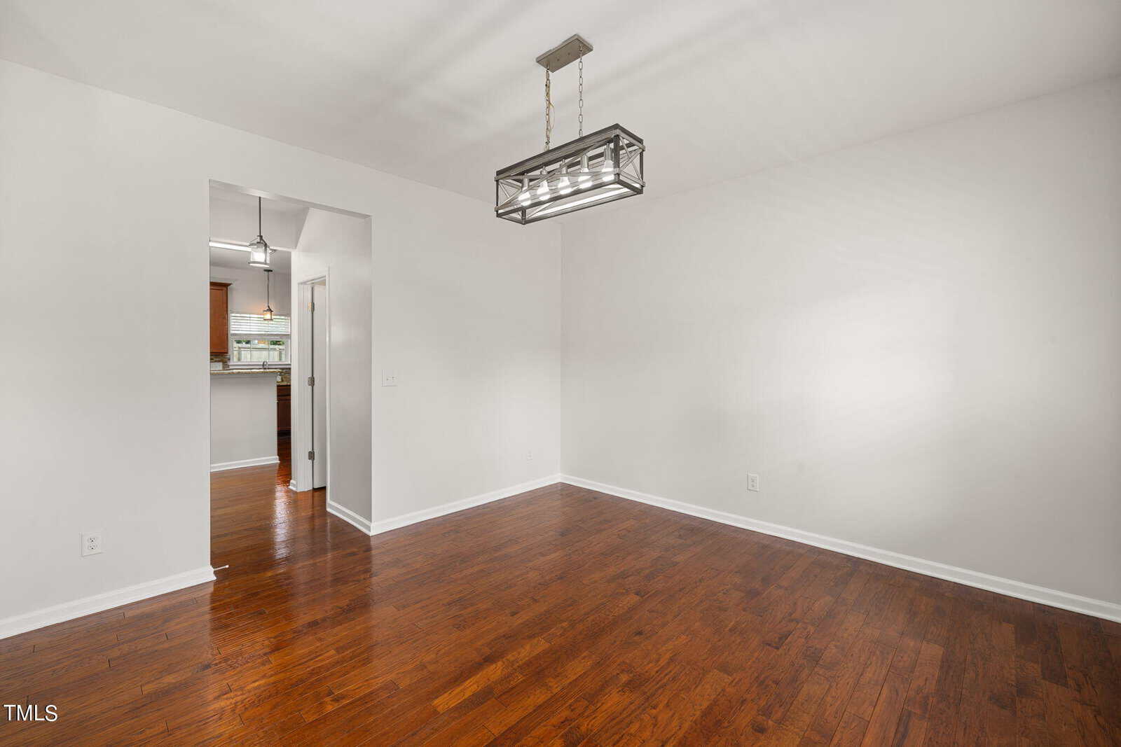 Empty white room with a wooden floor and a modern rectangular ceiling light fixture. There's an open doorway leading to a kitchen area with brown cabinets. The room is well-lit with natural light.