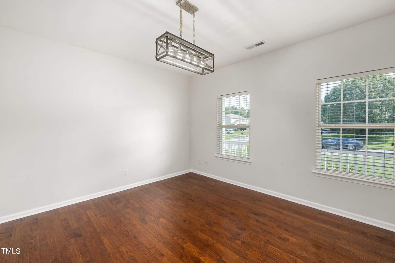 An empty room with white walls and hardwood floors, featuring a modern rectangular chandelier. Two windows with blinds are on one wall, overlooking a suburban street with greenery outside.