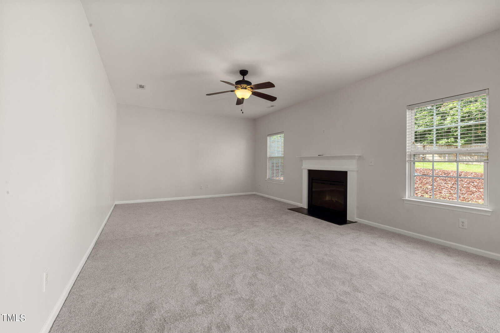 A spacious, empty living room with light gray carpet and white walls. A ceiling fan with a light hangs from the ceiling. The room has a fireplace beneath a small window, and a larger window provides natural light from outside.