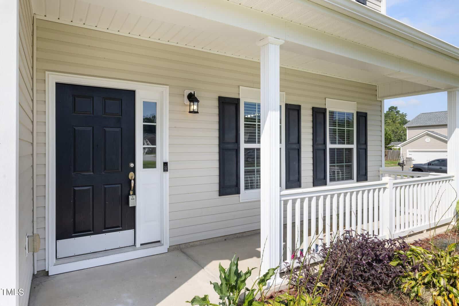 Front porch of a house with white railing and beige siding. Features a black front door with a lockbox, two windows with black shutters, and a lantern-style light. Small garden with green and purple plants lines the edge of the porch.