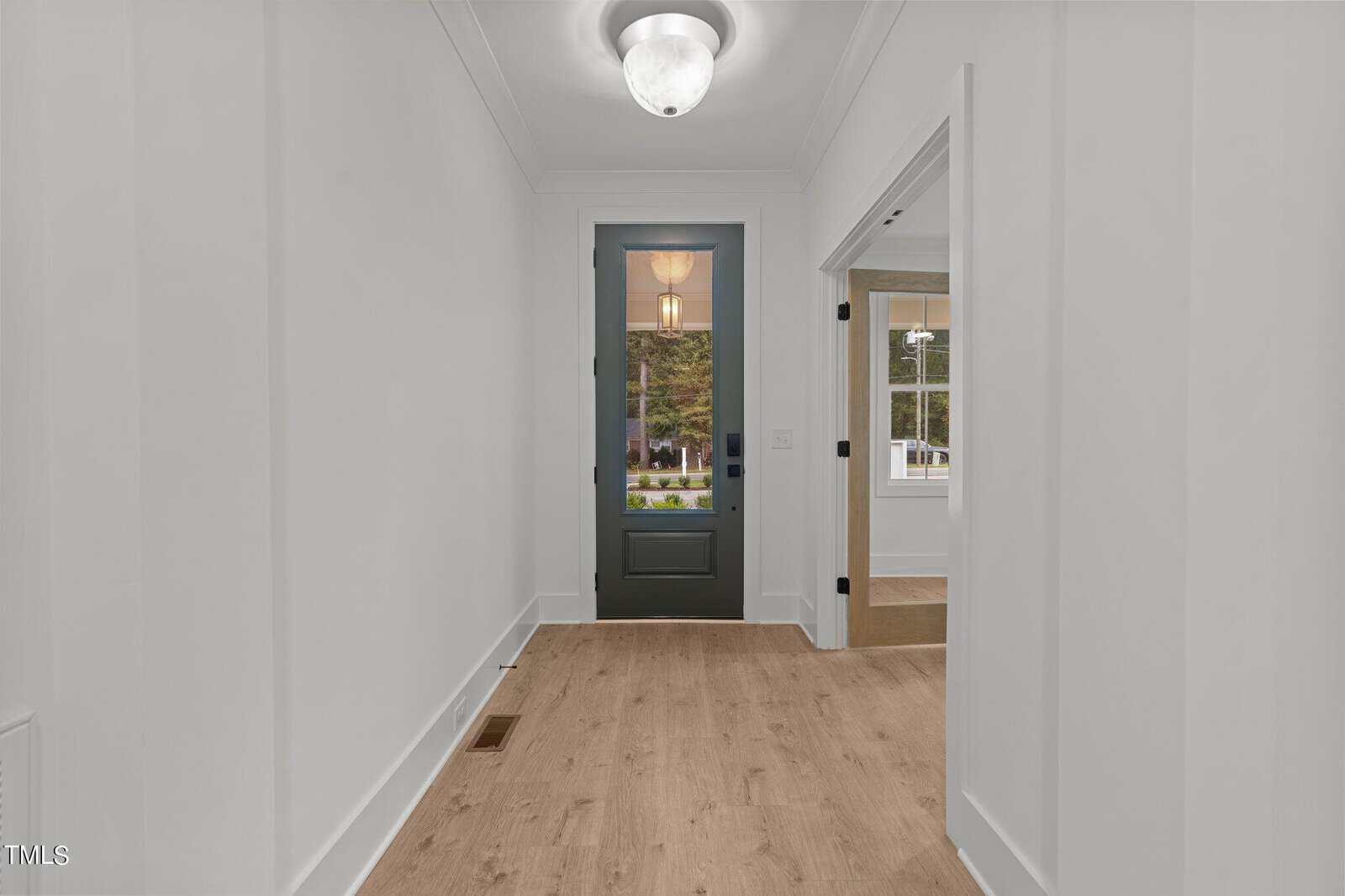 A bright hallway with light wood floors and white walls, leading to a glass-paned front door. A room with large windows is visible to the right, and a ceiling light fixture is overhead. Trees can be seen through the door.