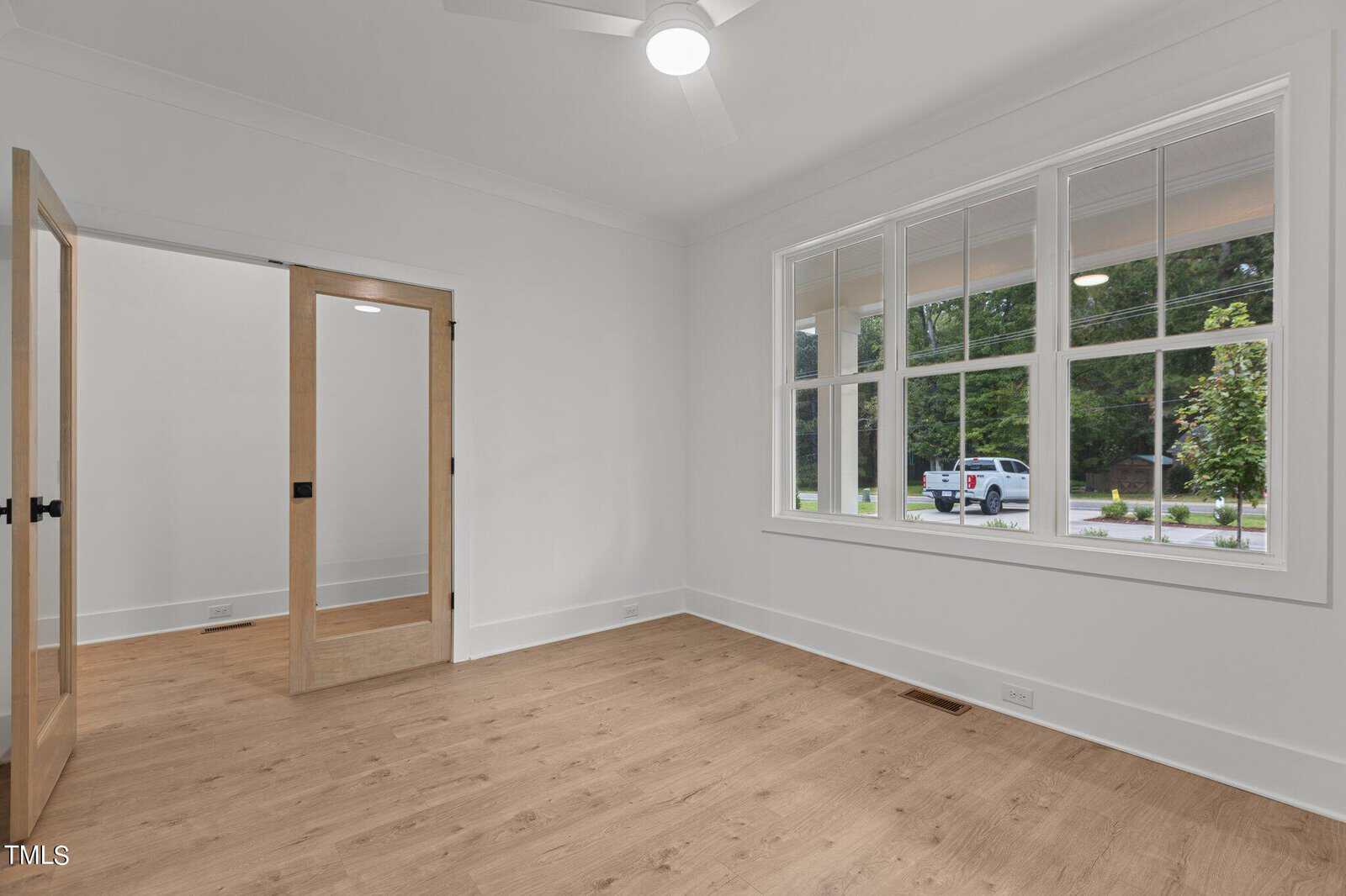 A bright, empty room with light wood floors, white walls, large windows, and glass double doors, offering a view of a parked car and greenery outside.