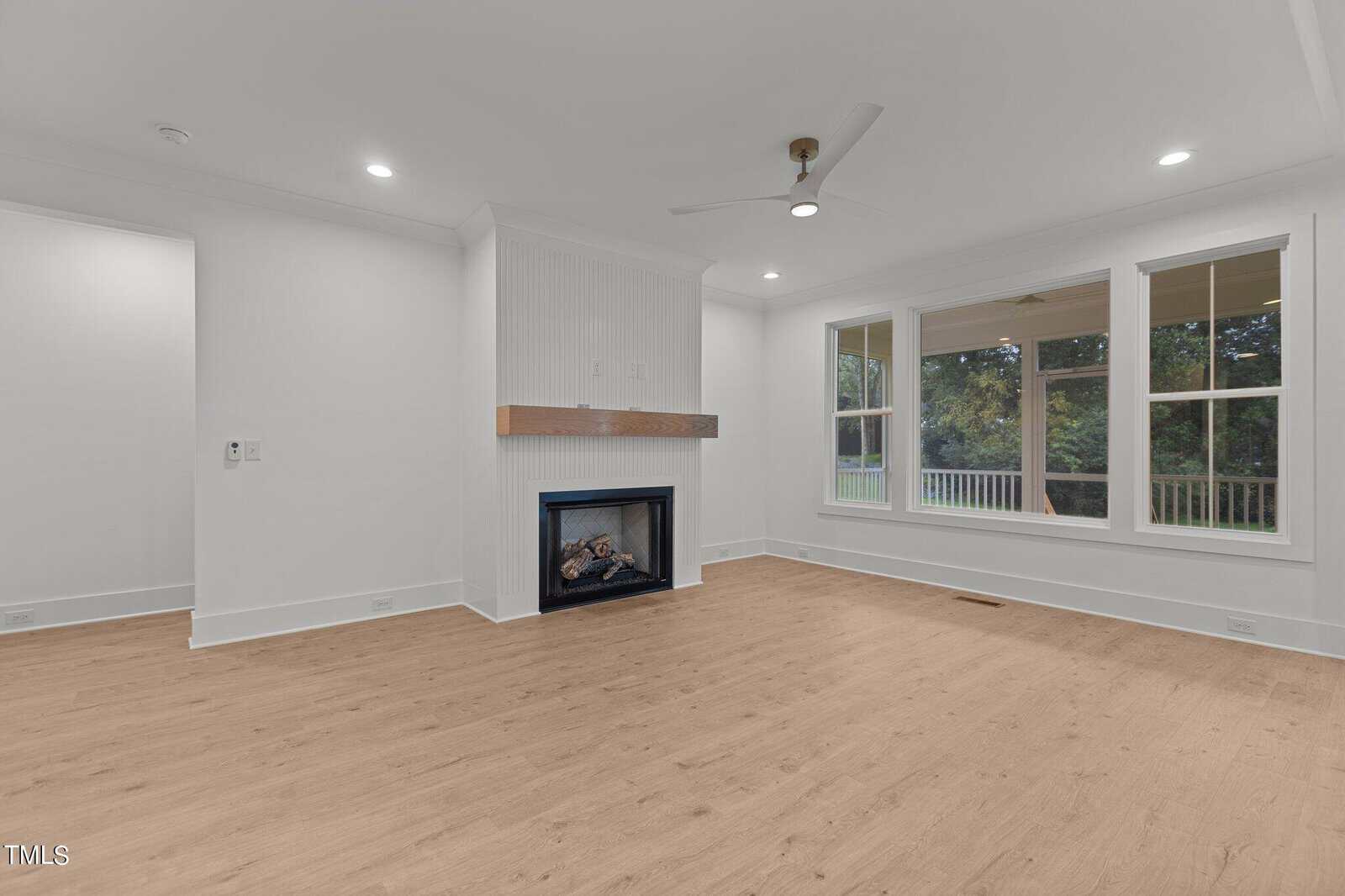 A bright, empty living room with light wood floors, white walls, a modern ceiling fan, a fireplace with a wood mantel, and large windows looking out to green trees.