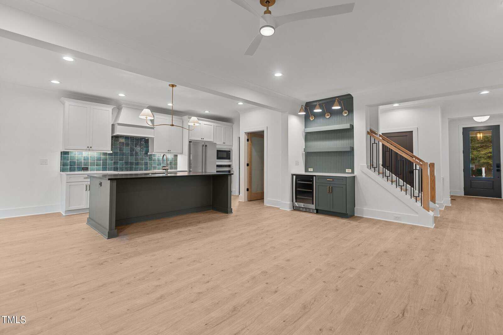 Modern open-concept kitchen and living area with light wood floors, white walls, an island with dark cabinetry, teal tile backsplash, pendant lighting, and a staircase leading upstairs. A glass door is visible in the background.