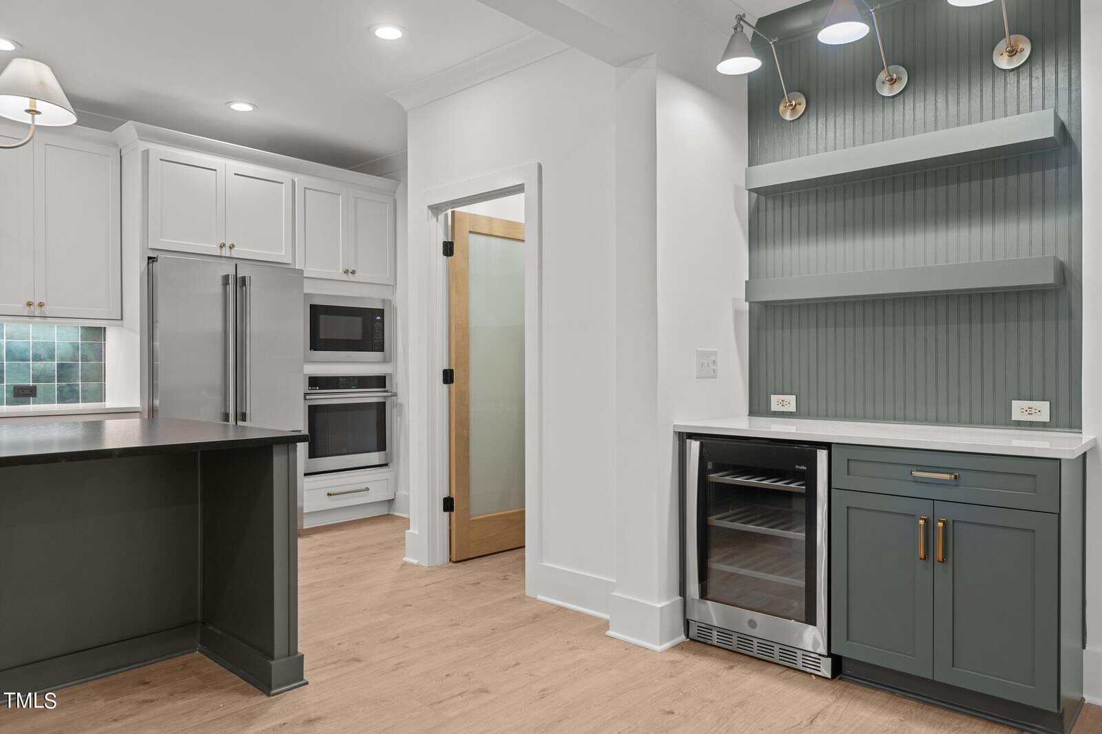Modern kitchen with white cabinets, stainless steel appliances, light wood floors, a built-in oven and microwave, green open shelves, wine fridge, and a glass-paned door. Neutral colors and clean lines create a contemporary look.