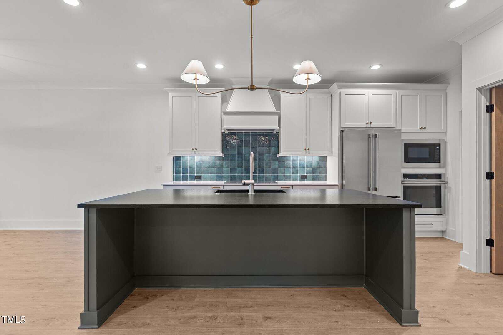 Modern kitchen with white cabinets, a large dark island with a built-in sink, light wood flooring, stainless steel appliances, blue tiled backsplash, and a double chandelier above the island.