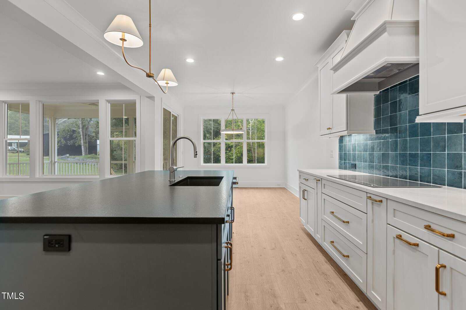 Modern kitchen with a large black island, white cabinets, gold hardware, blue tile backsplash, pendant lights, wood flooring, and large windows overlooking a green yard.