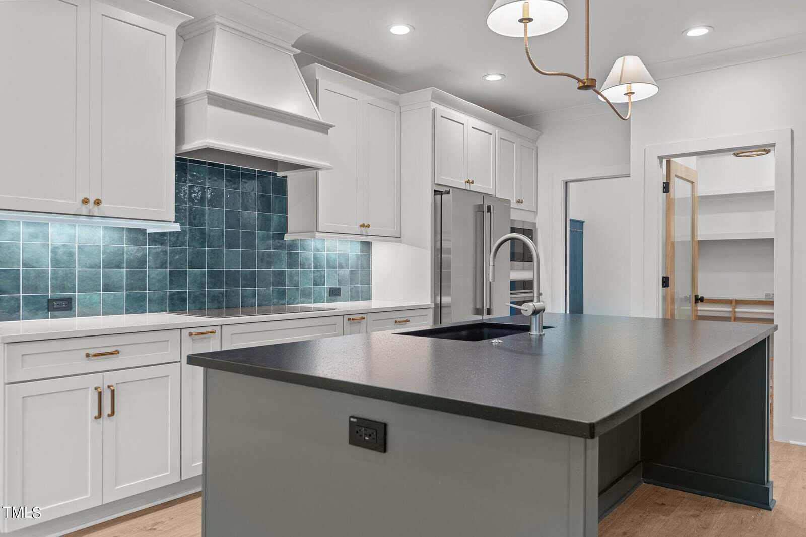 Modern kitchen with white cabinets, a black countertop island with a sink, stainless steel appliances, blue tile backsplash, wood flooring, and brass light fixtures. A pantry with glass door is visible in the background.