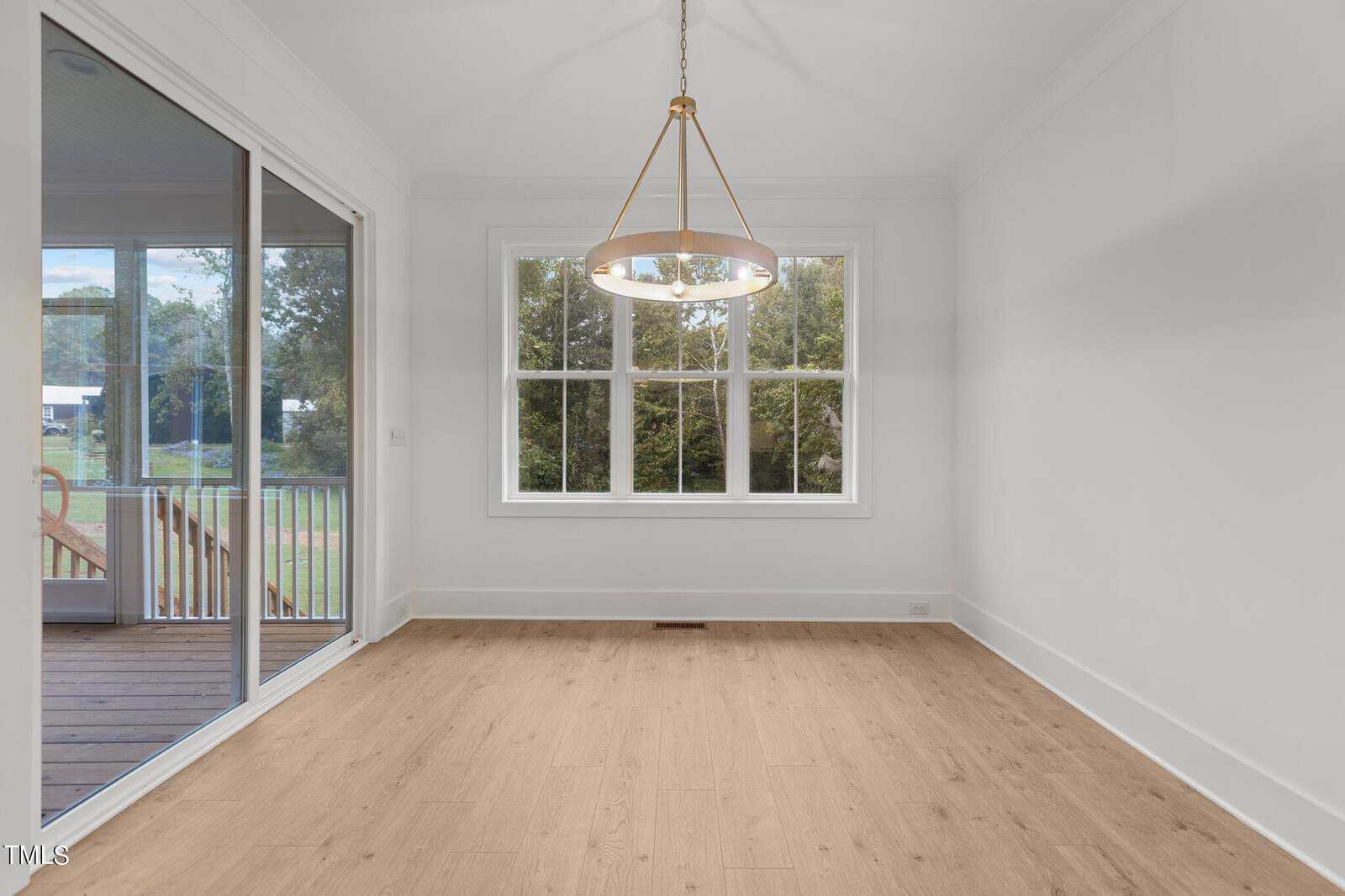 Bright, empty room with light wood floors, a large window overlooking trees, a modern round chandelier, and glass doors leading to a porch with stairs outside.