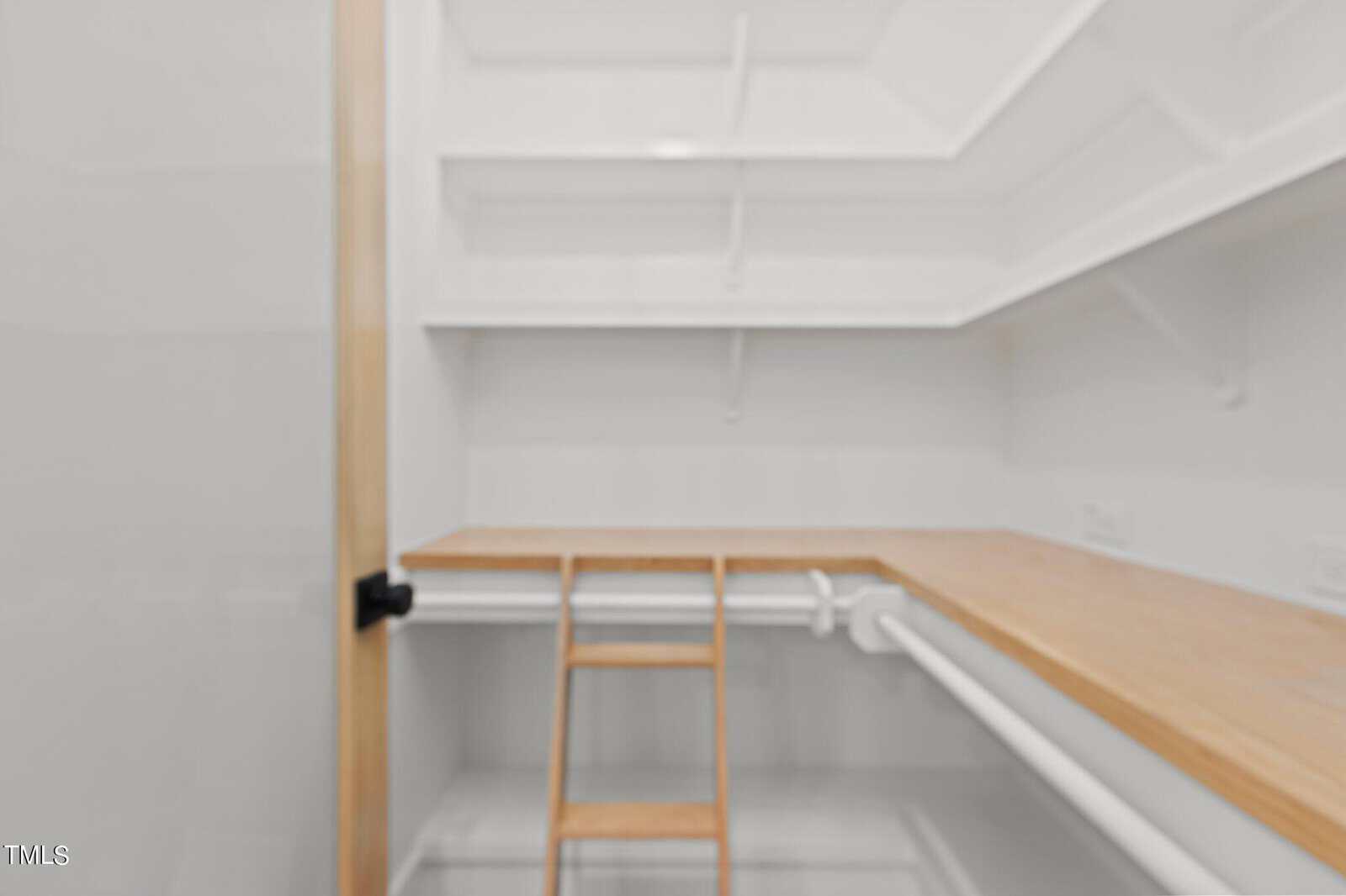 A walk-in closet with wooden shelves and a wooden countertop, featuring a small wooden ladder, white walls, and empty hanging rods. The space appears clean and well-organized.