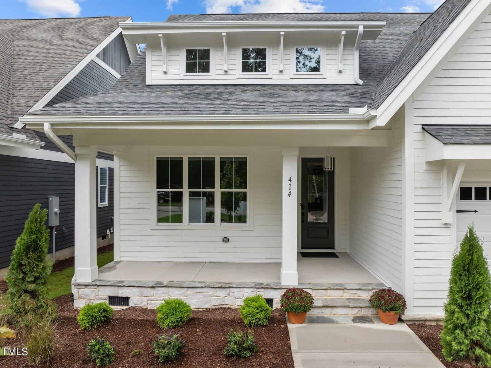 A modern white house with a covered front porch, dark green door, large window, gray shingle roof, stone base, and neatly landscaped garden with shrubs and potted plants.