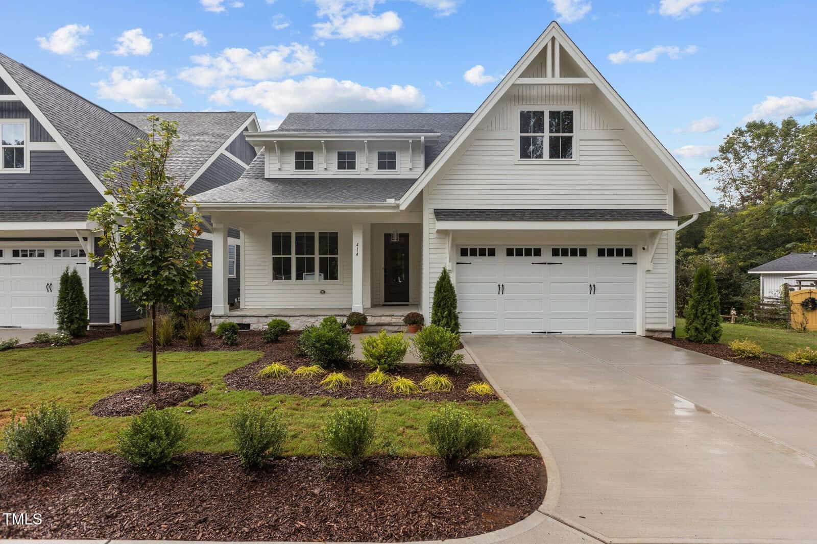 A modern two-story white house with a front porch, gabled roof, and attached two-car garage. The yard is landscaped with small shrubs, a tree, and a concrete driveway under a partly cloudy sky.