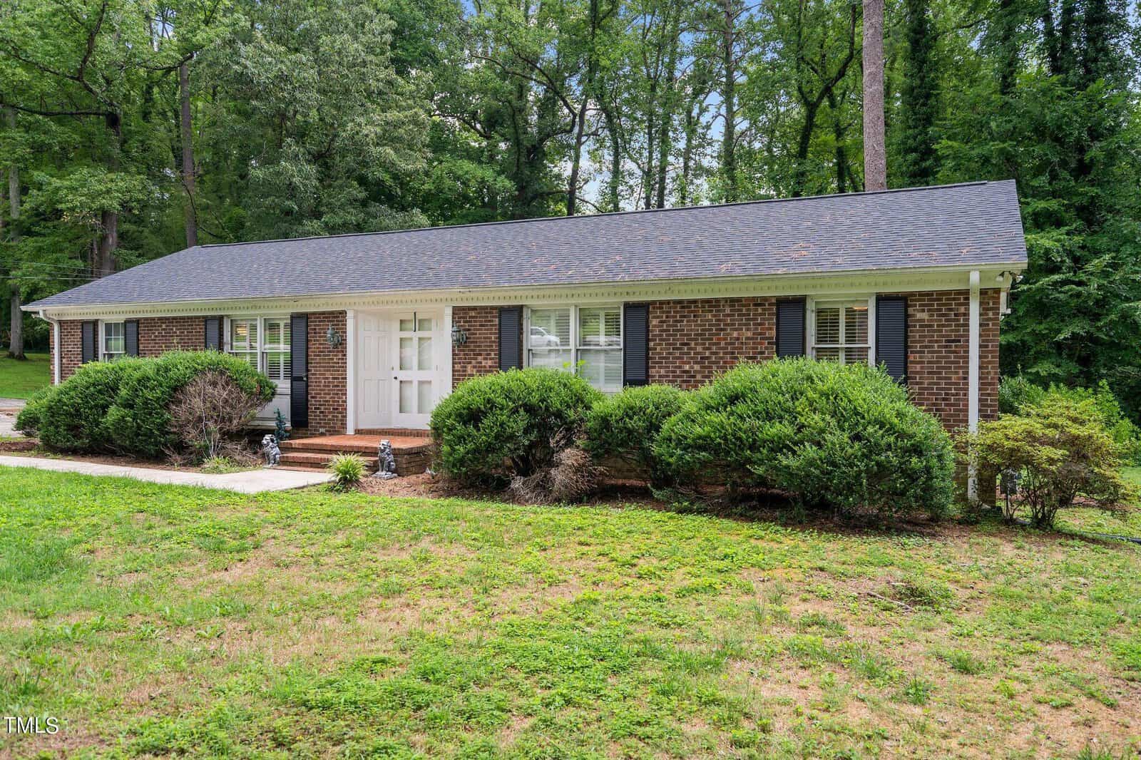 A single-story brick house at 1707 Wendover Road, with dark shutters and a gray shingled roof, is surrounded by greenery. The front door is framed by two large windows and flanked by bushes, with a small set of stairs leading up to it. Tall trees are visible in the background of this Henderson home.