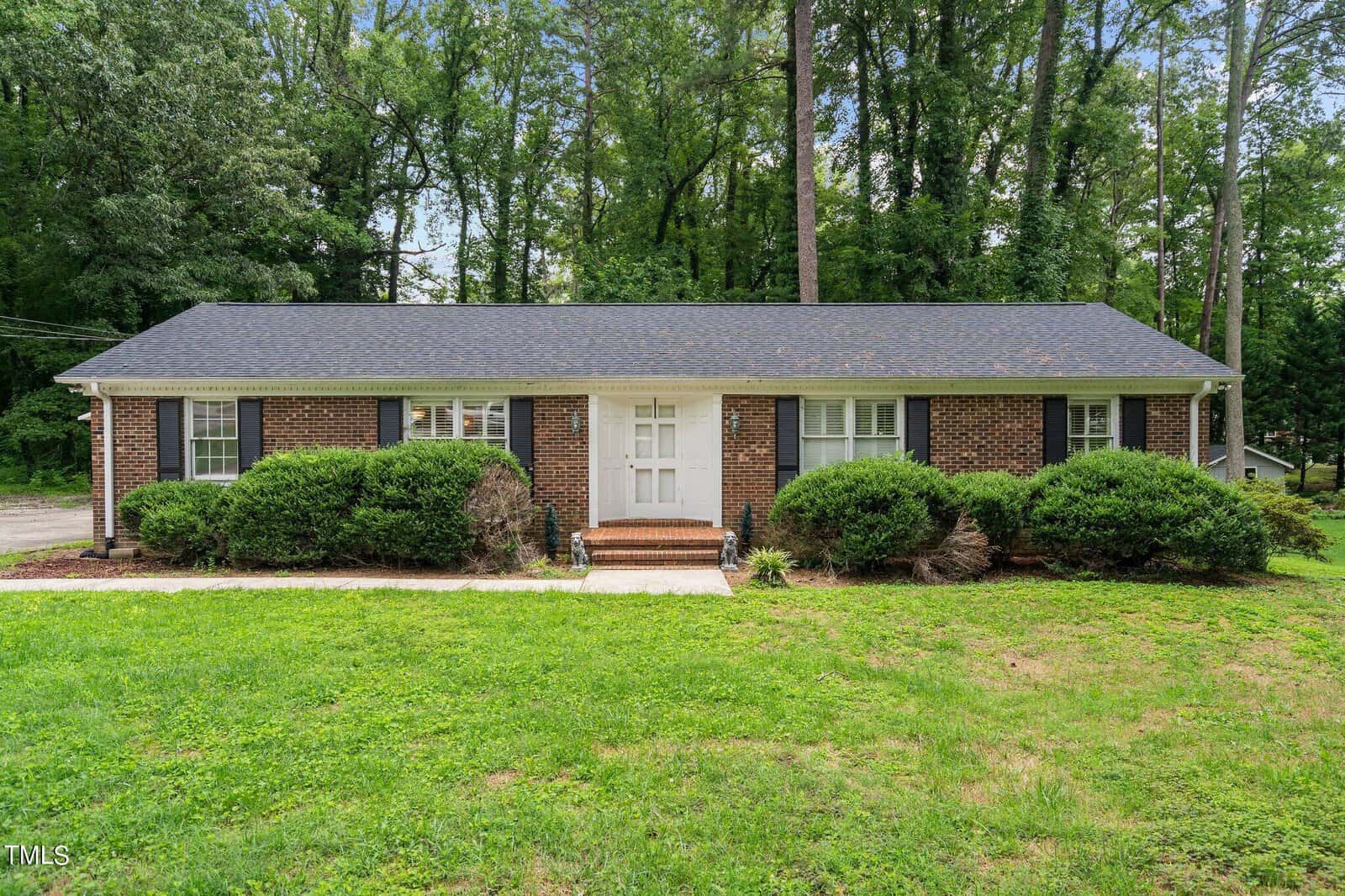 A single-story brick house with a dark shingle roof, white front door, and black shutters. The house is surrounded by neatly trimmed bushes and set against a backdrop of tall, green trees. A small lawn is visible in the foreground with a pathway leading to the entrance.