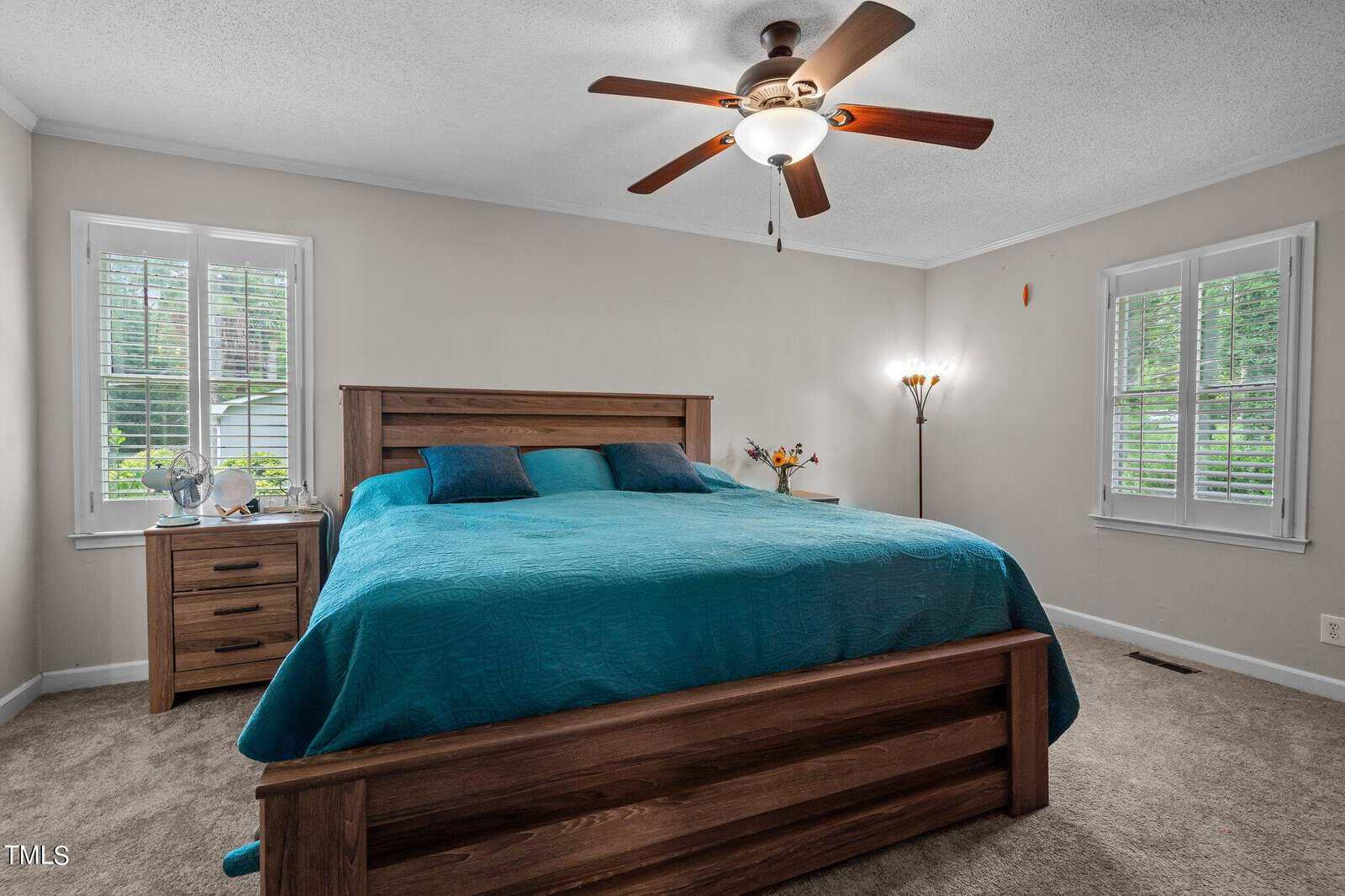 A bedroom features a large wooden bed with a teal comforter and pillows. On one side of the bed is a wooden nightstand with a lamp, and on the other side, a floor lamp with multiple lights. The room has windows with white trim providing natural light.