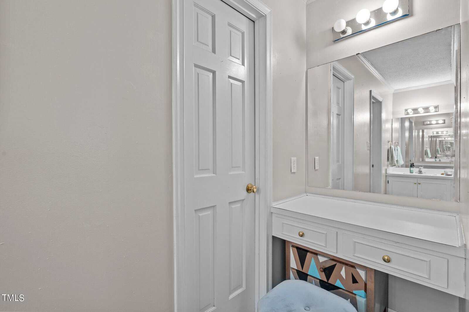 A white-walled room featuring a door to the left and a vanity area with a large mirror on the right. The vanity has a drawer with golden knob and a small blue cushioned stool underneath. There are several light bulbs above the mirror.