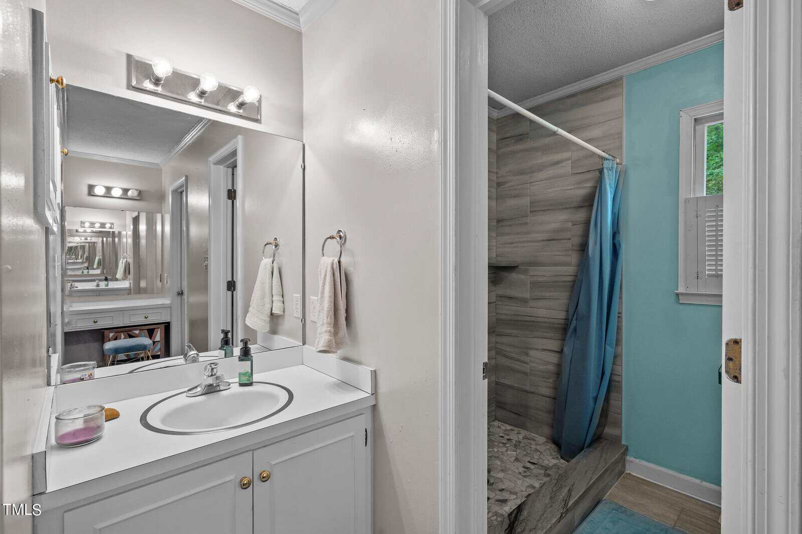 A bathroom with a white vanity and sink, a mirror with lights above, and various toiletries on the counter. Adjacent is a shower with gray tiling and a blue shower curtain. There's a small window with blinds in the shower area.