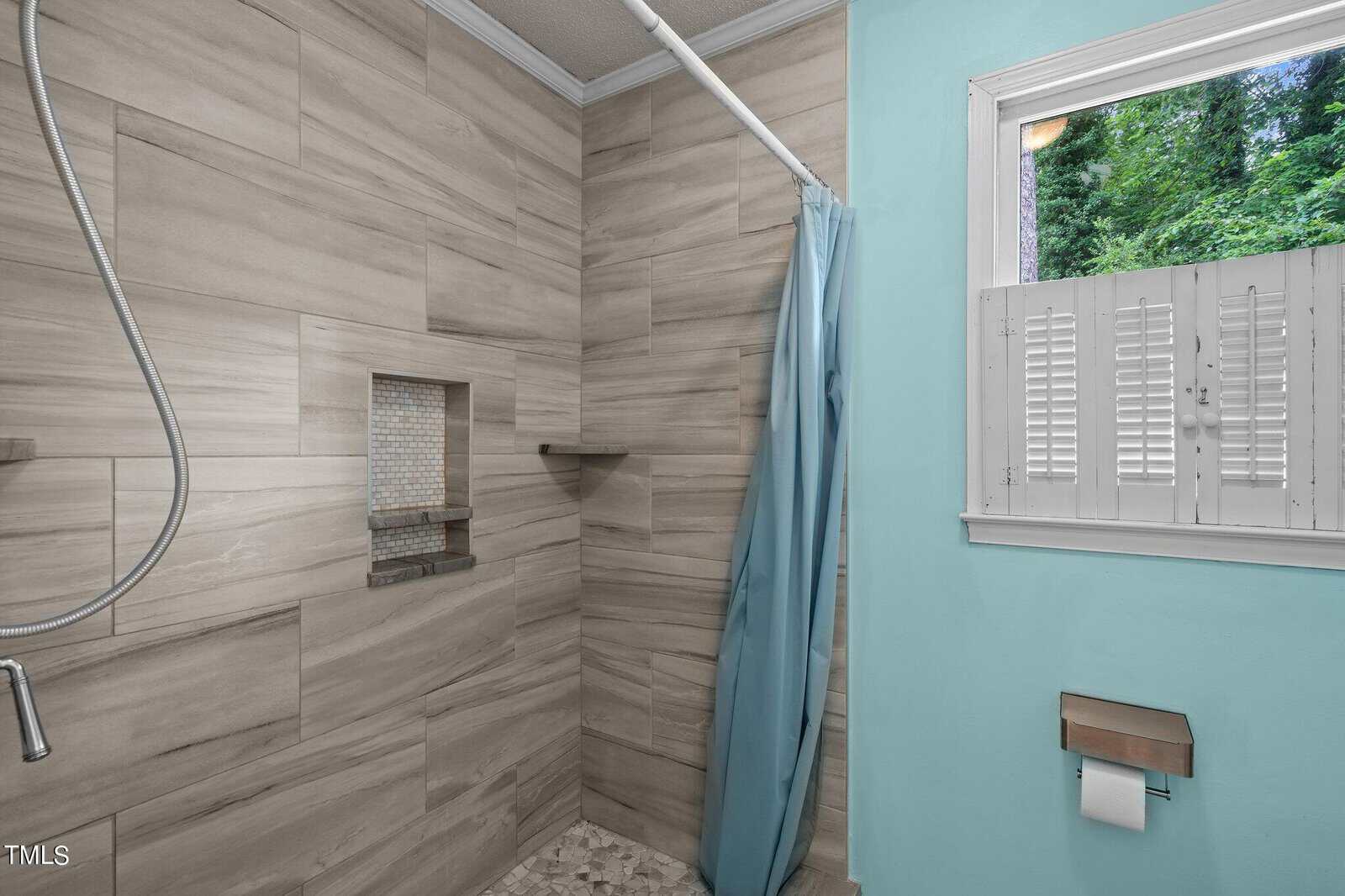 A modern bathroom with beige tiled walls and a shower area featuring a blue curtain, a built-in niche with mosaic tiles, and a handheld showerhead. Adjacent is a window with white shutters, and below it, a toilet paper holder attached to a light turquoise wall.