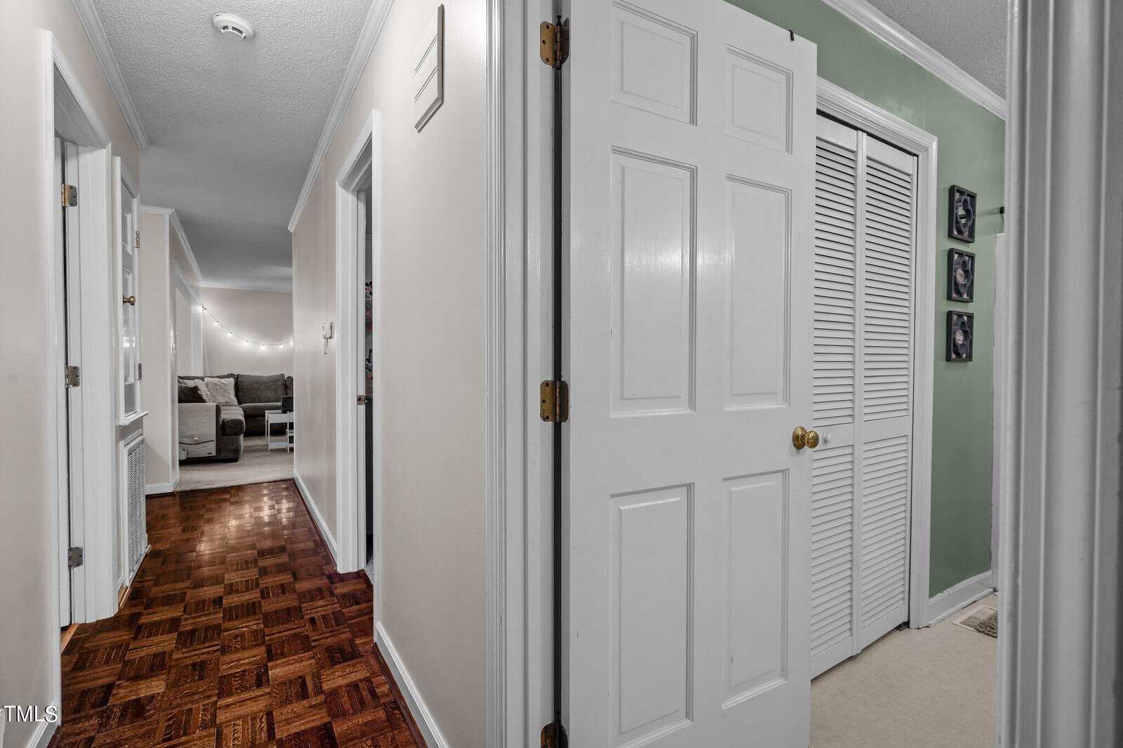 A hallway with wood parquet flooring leads to a living room where a sectional couch and string lights are visible. On the right, there is a partially open door revealing a room with green walls and a louvered closet door. White walls and trim accent the space.