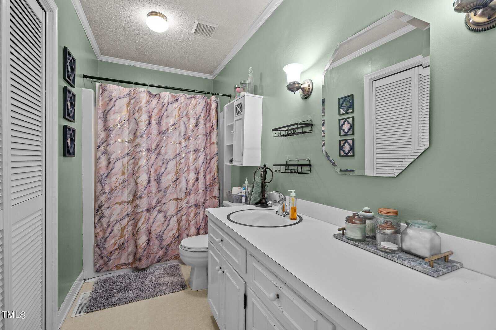 A bathroom with green walls featuring a large white countertop with two sinks and various toiletries. There is a white cabinet above the counter, a toilet beside it, and a shower with a pink and white marbled curtain. A mirror and sconces are above the sinks.