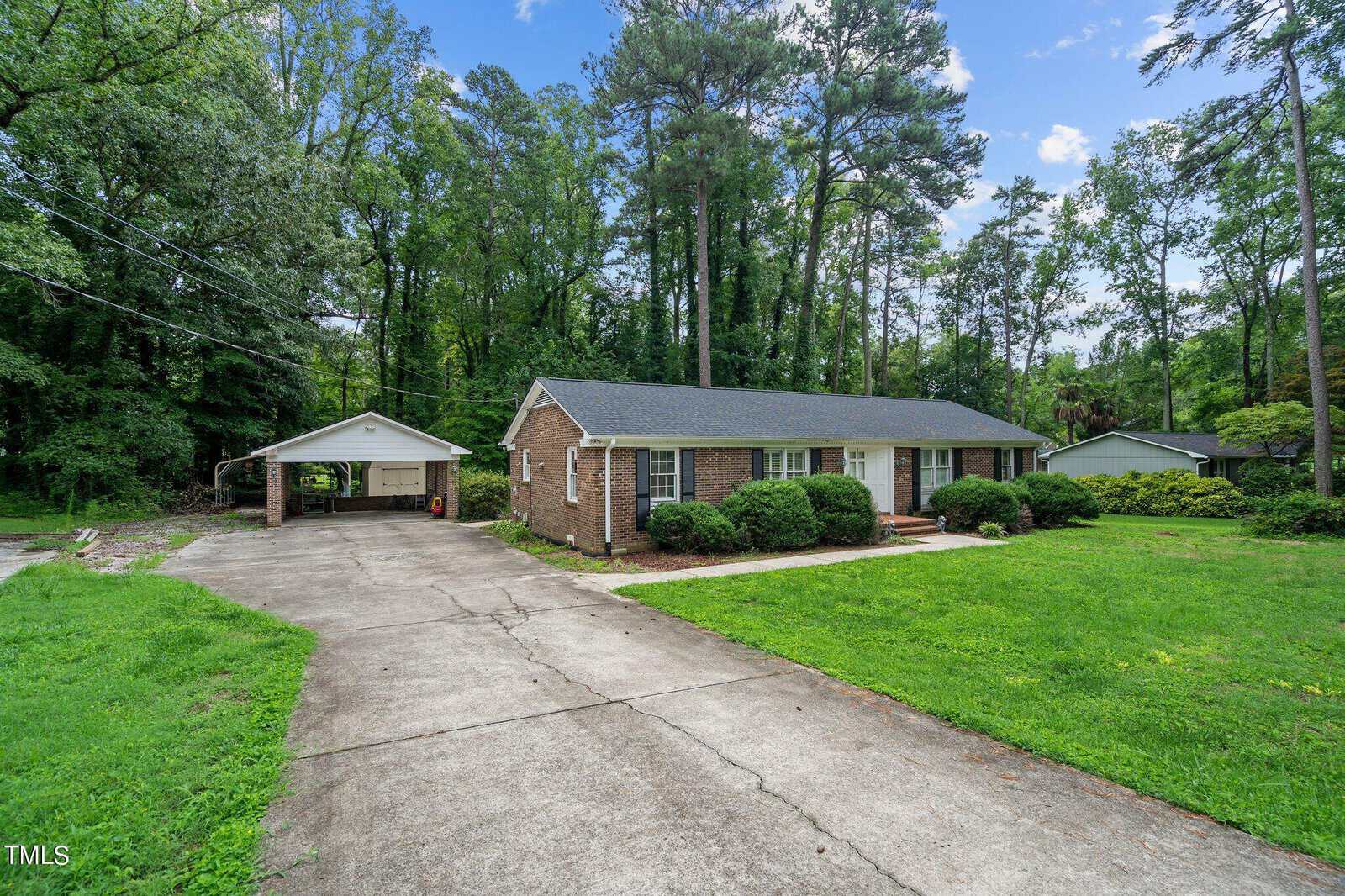 A single-story brick house with a dark roof sits among tall trees. The front yard has green grass, and a concrete driveway leads to a carport. The house has white-framed windows and a small front porch. The surrounding area is wooded, providing ample shade.