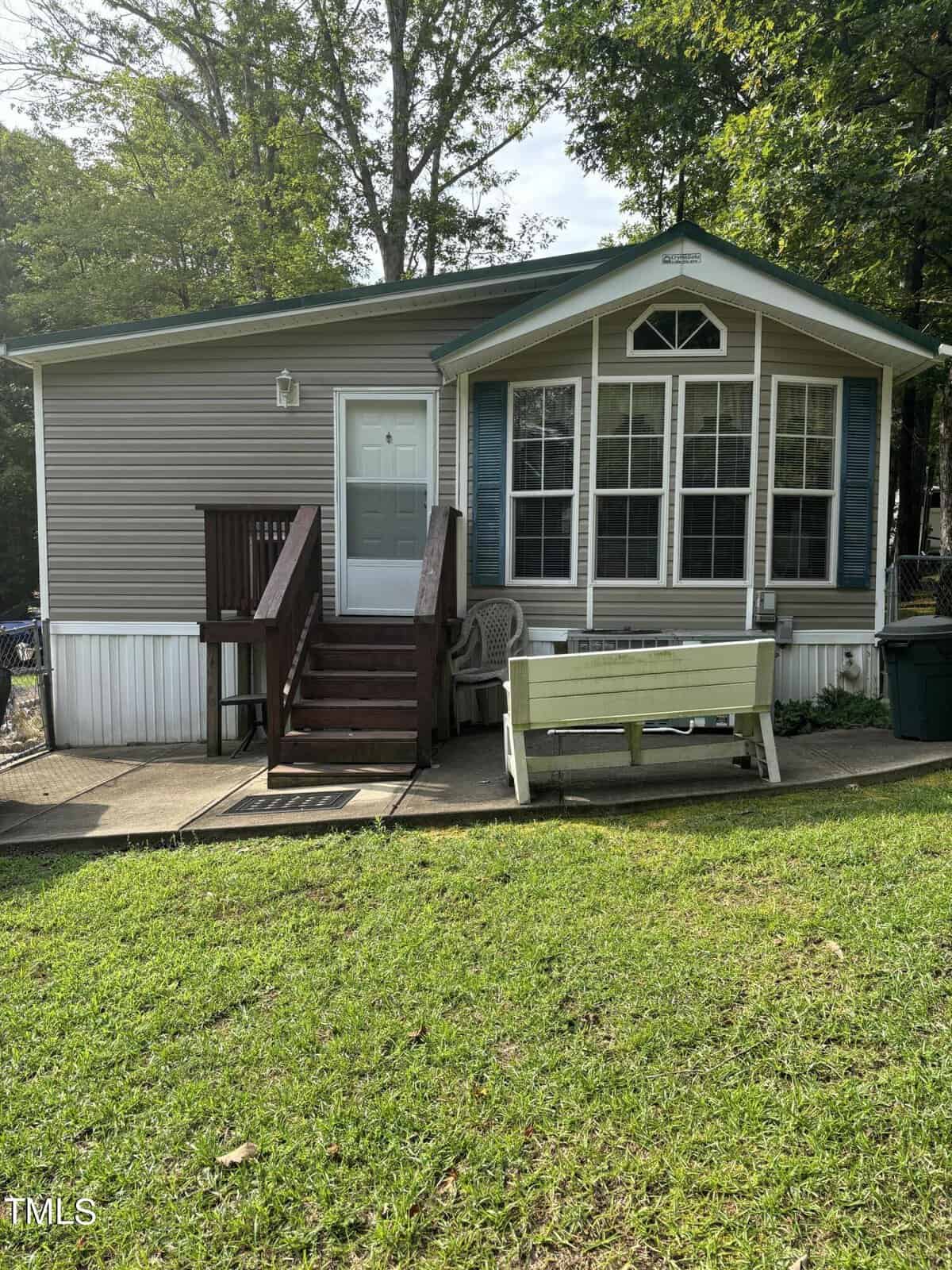 A small beige manufactured home with green trim located at 104-106 Catoose Drive in Louisburg, featuring a front porch with stairs, a white bench, two chairs, and large windows. The house sits on a grassy yard with trees in the background.