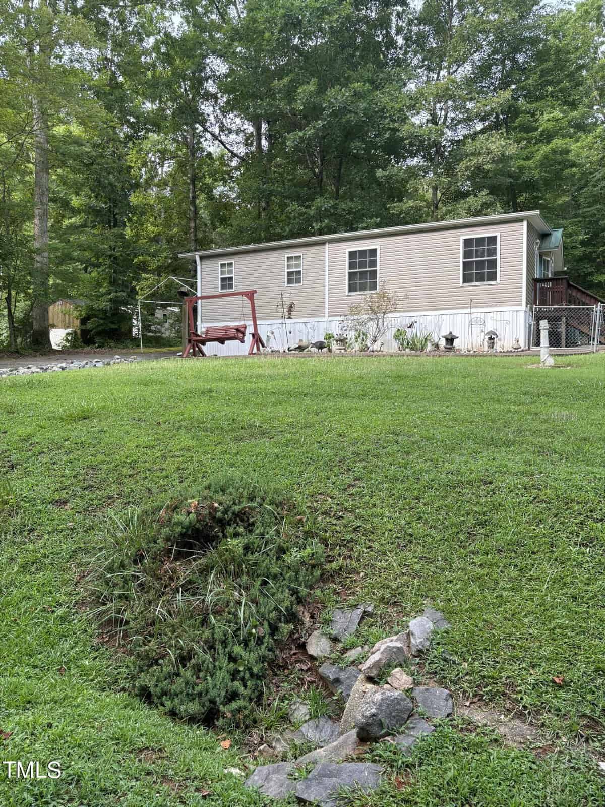 A beige mobile home sits on a grassy lawn with a red porch swing and small garden in front. The yard is bordered by trees, and rocks line a depression in the grass. A fenced area is visible on the right side.