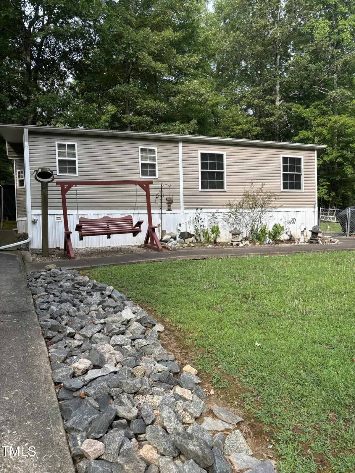 A beige and white mobile home with three windows, a covered porch, and a red wooden swing in the yard. Rock landscaping lines the driveway, and there are trees in the background.