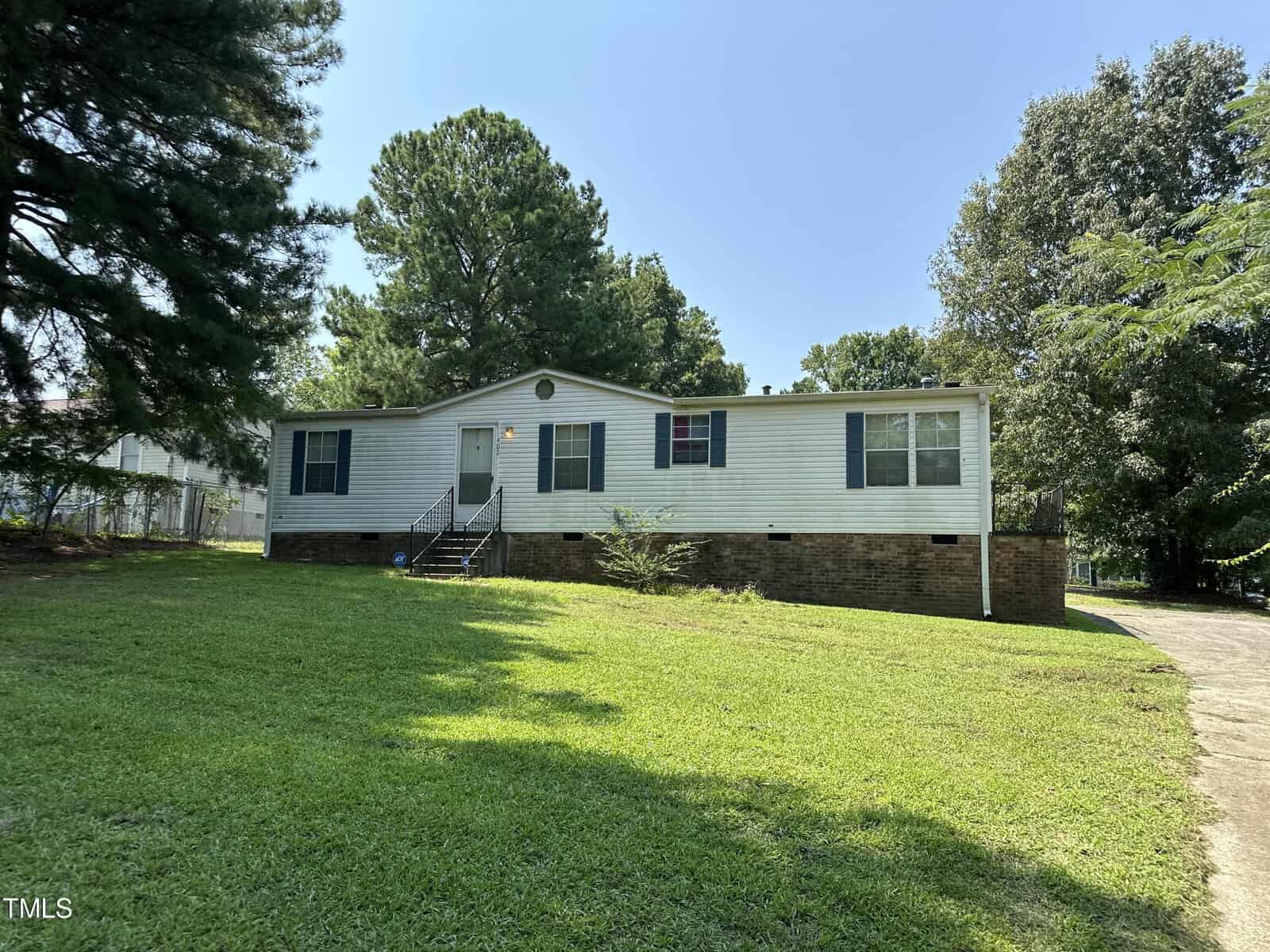 Nestled at 200 Pecan Street in Creedmoor, a white single-story house with blue shutters and a brick foundation sits surrounded by trees on a sunny day. The yard is grassy, and a small set of stairs leads to the welcoming front door.