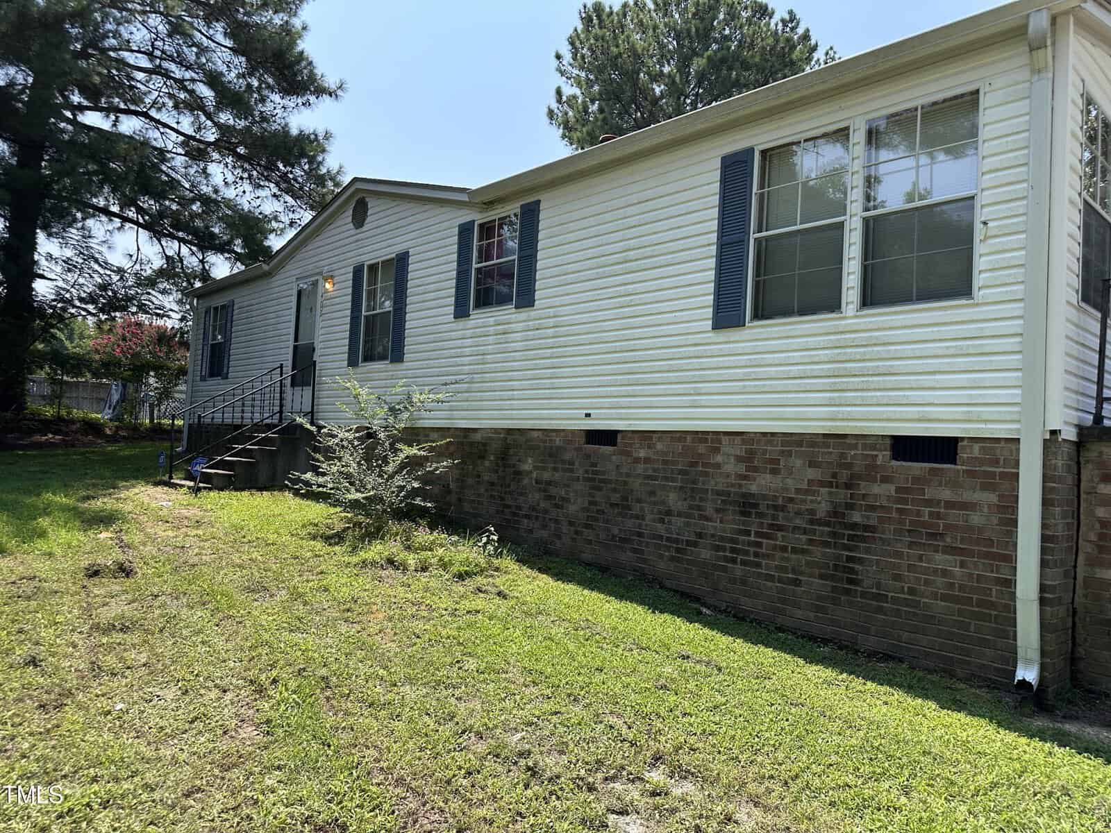 A single-story house with white siding and dark shutters sits elevated on a brick foundation. It has a grassy yard with a small tree next to the front steps. Tall trees border the property in the background.