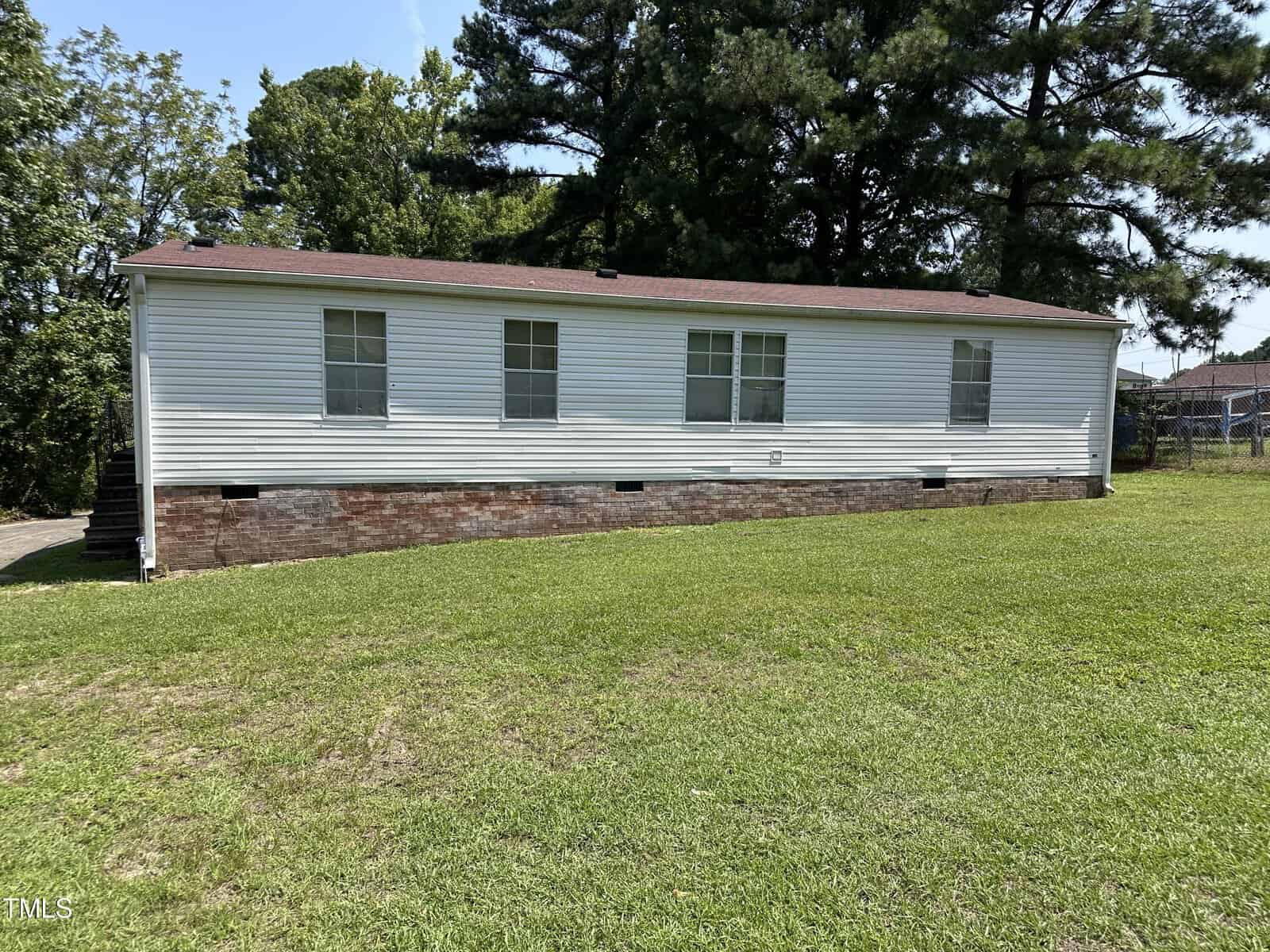A white single-story mobile home with a red-brown shingled roof sits on a grassy lawn. The house has several windows and brick skirting, surrounded by trees under a clear blue sky.