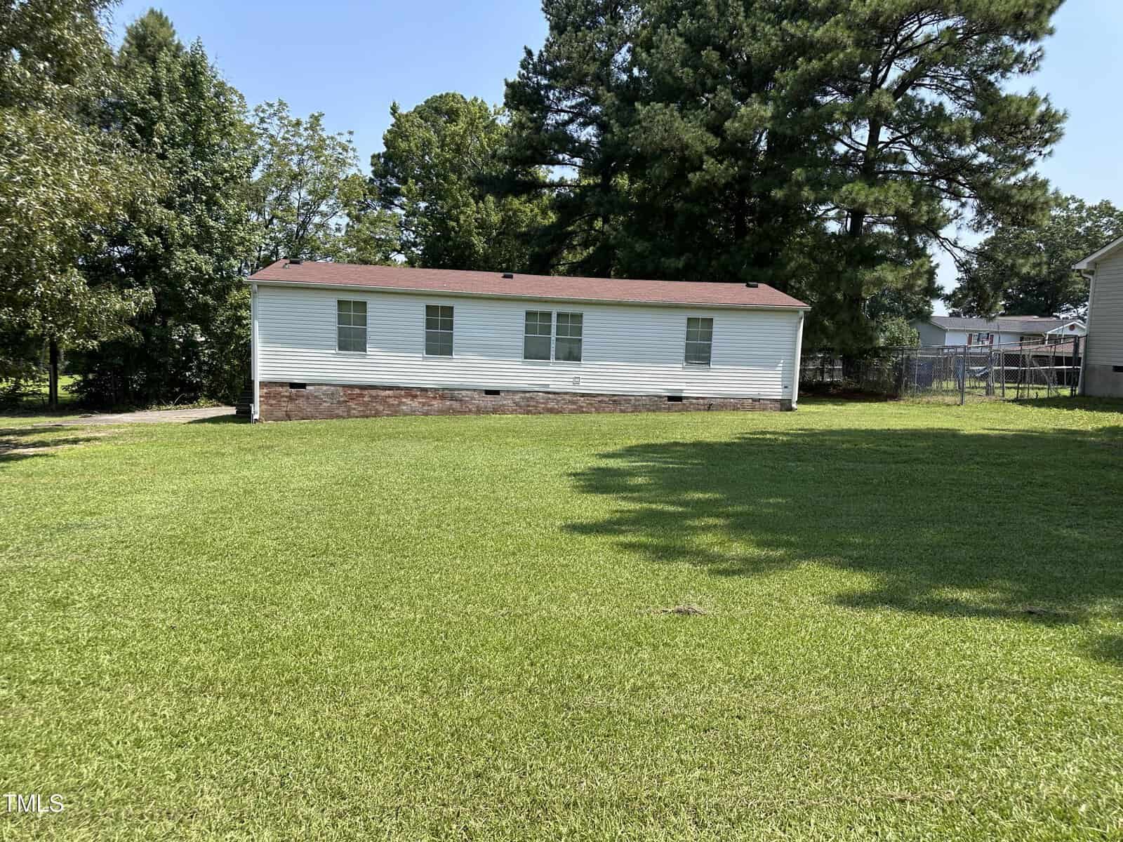 A small white mobile home with a red-brown roof sits on a large, neatly trimmed grassy lawn. Tall green trees surround the house, and a fence is visible on the right side.