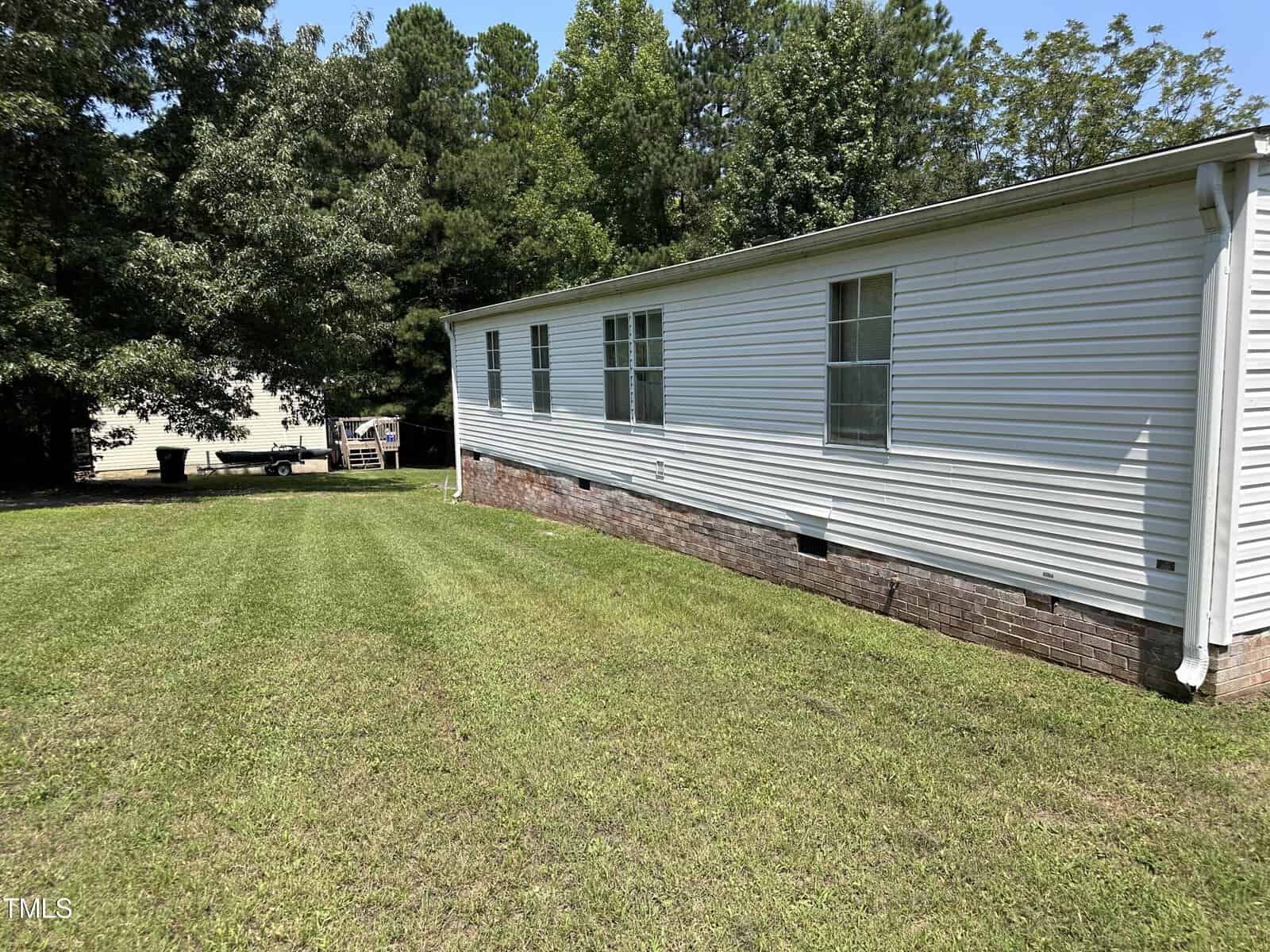 A single-story white mobile home with several windows is situated on a grassy lawn. In the background, there are dense trees and a small structure partially visible. The sky is clear and sunny.