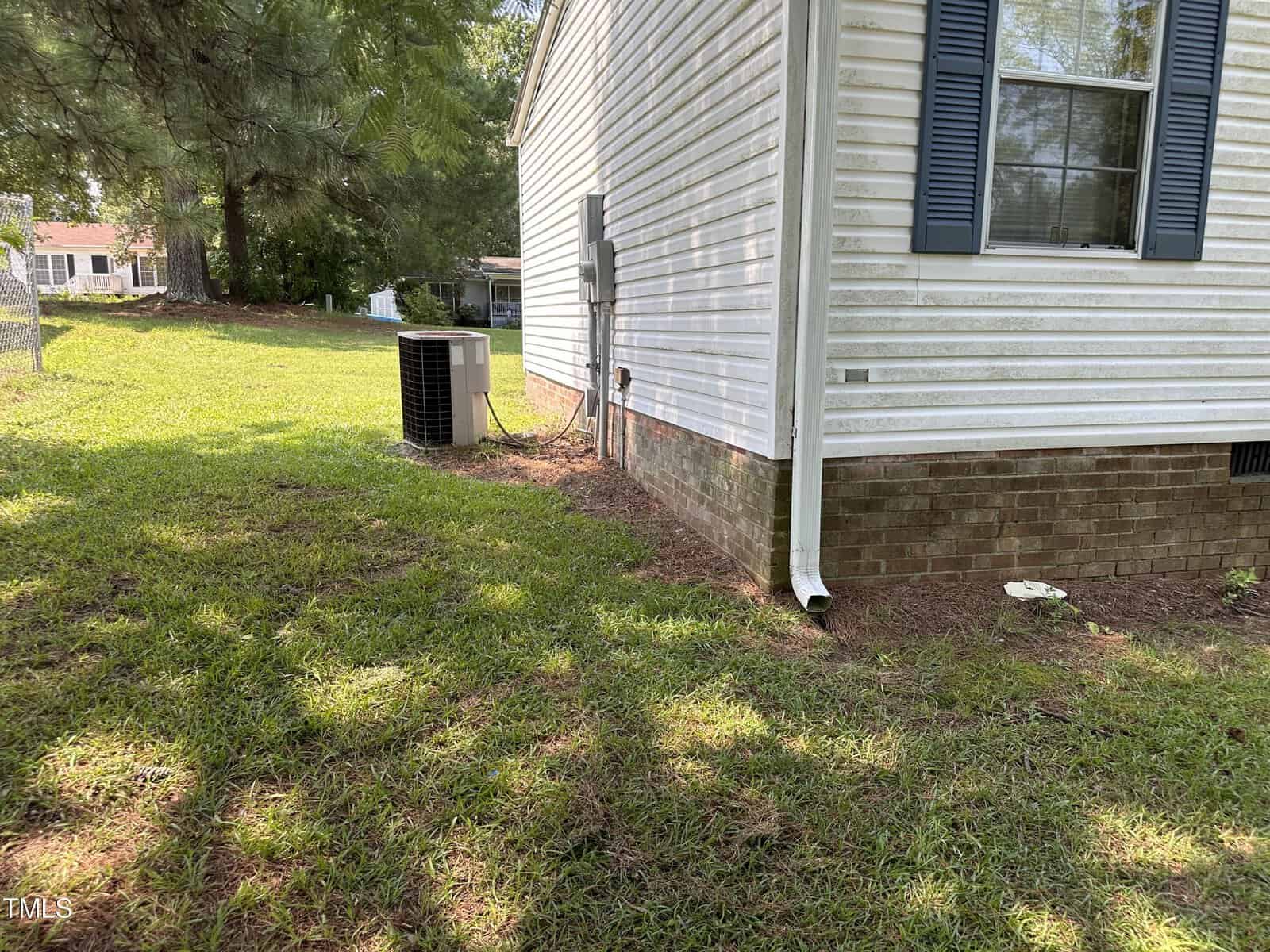 Side view of a white house with brick foundation, featuring a blue shuttered window and a downspout. An air conditioning unit is visible by the wall. The yard is grassy, bordered by trees and a wire fence.