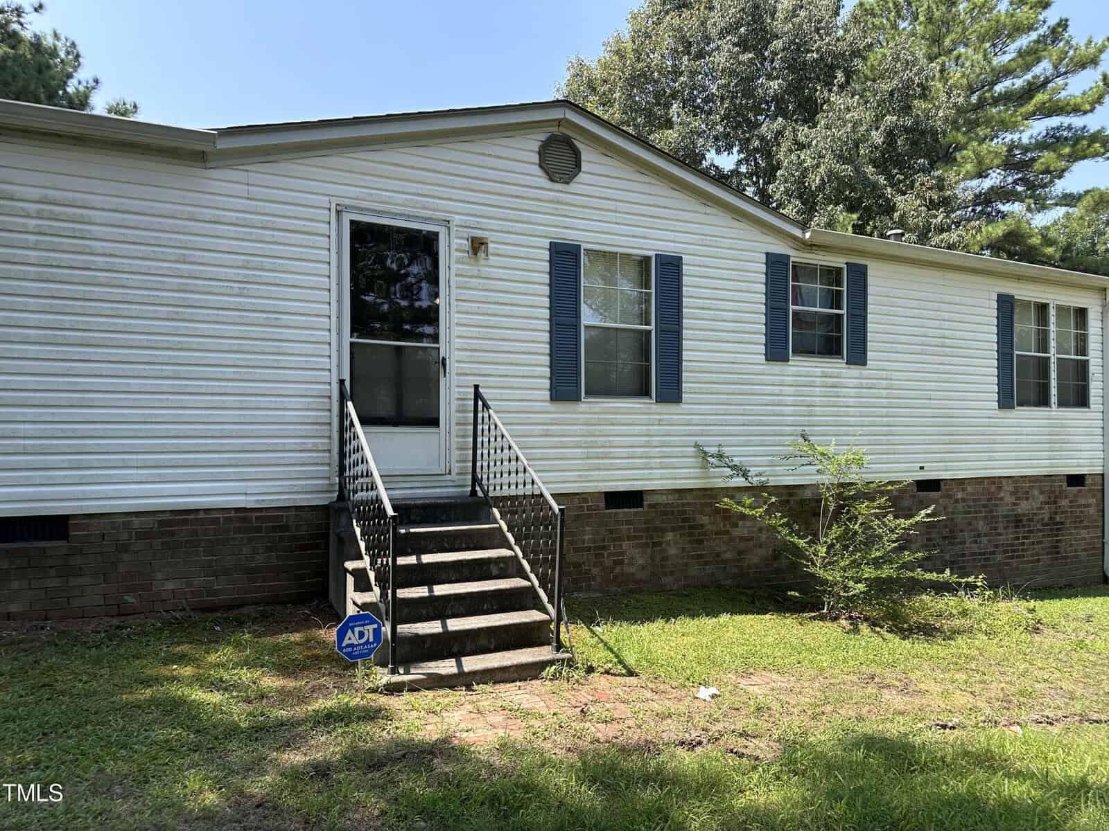 A white mobile home with blue shutters, a front door with a small set of stairs, and a brick foundation. The yard is grassy with a few small bushes. An ADT security sign is visible near the steps. Trees are in the background.