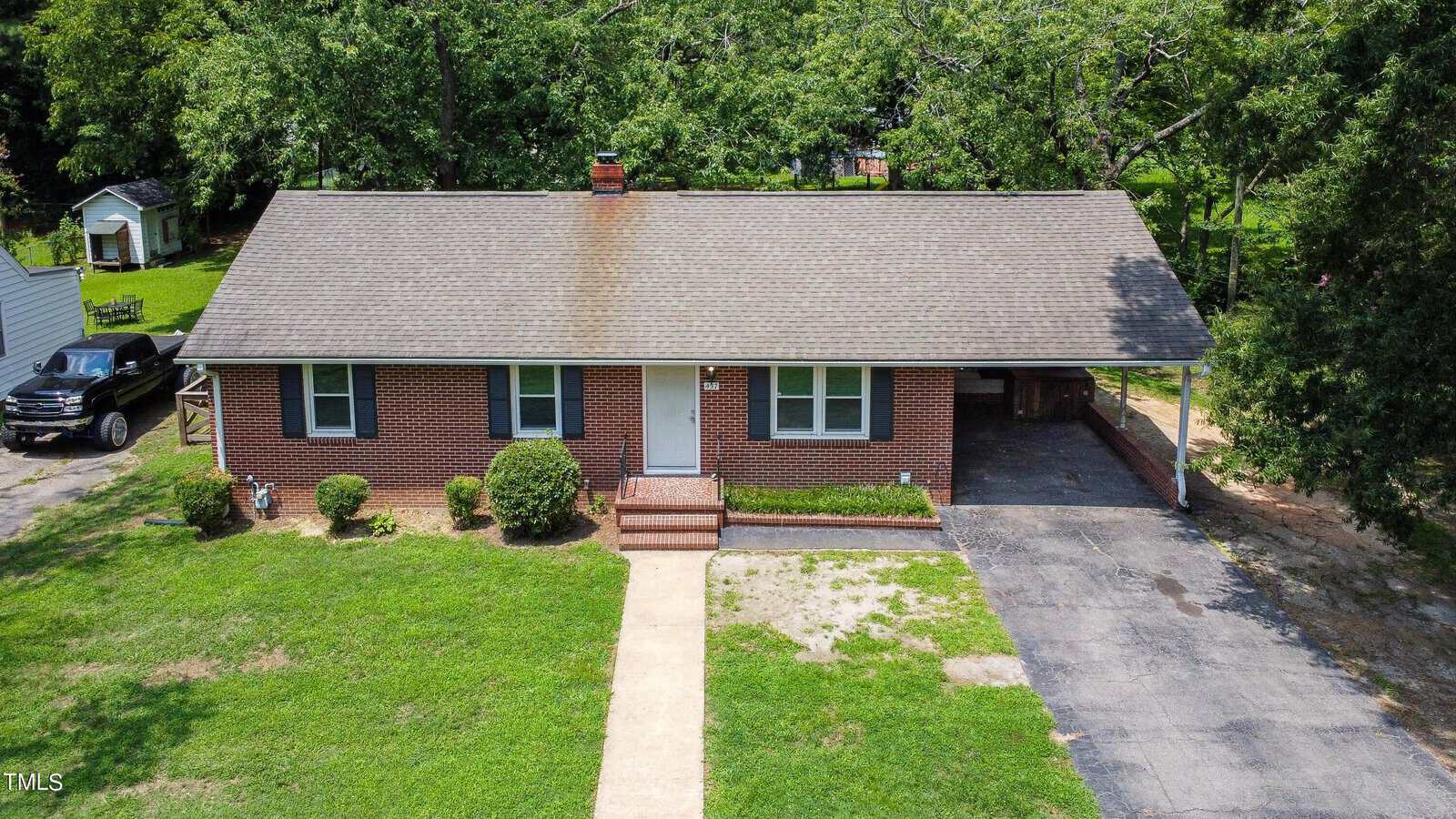 A single-story brick house at 437 Norvell Street, Henderson, features a gable roof, central front door, white-framed windows, and a carport with driveway. Trimmed bushes line the small porch, while green lawn and mature trees surround the home.