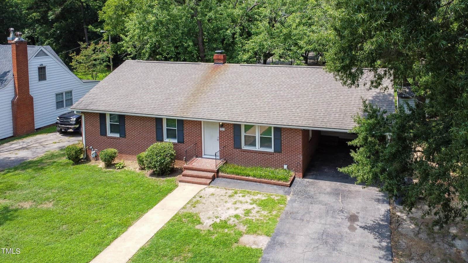 A single-story red brick house with a gray shingled roof, central chimney, and white trim. The home has a small front porch, carport on the right, driveway, and a lawn with shrubs; surrounded by trees and neighboring homes.