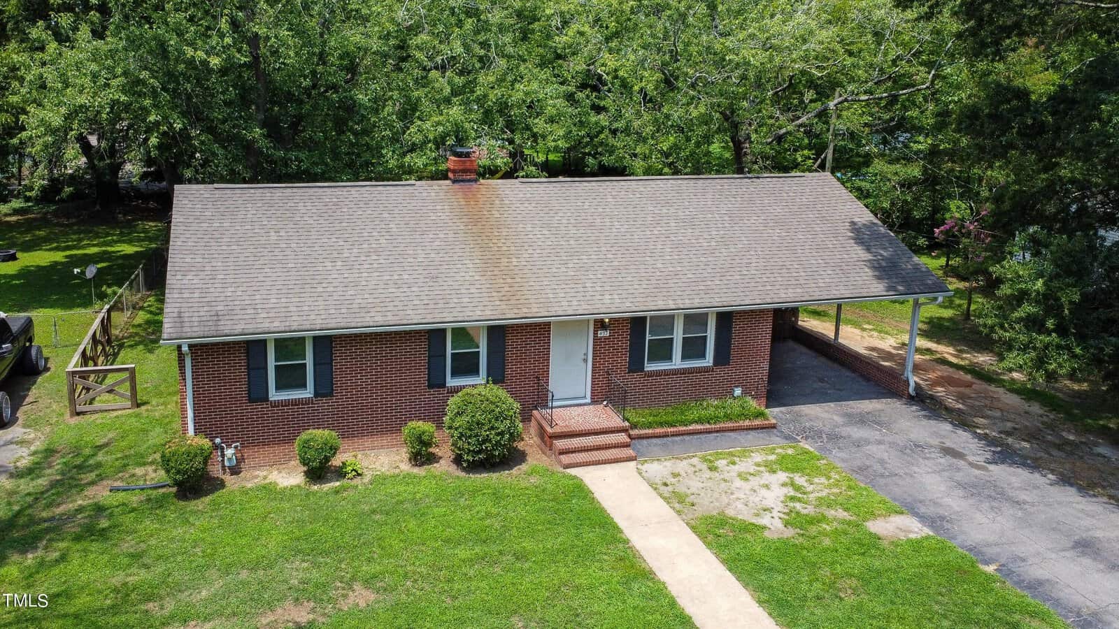 Aerial view of a single-story brick house with a shingled roof, central chimney, front steps, small shrubs, and an attached carport. The house sits on a grassy lawn with trees in the background.