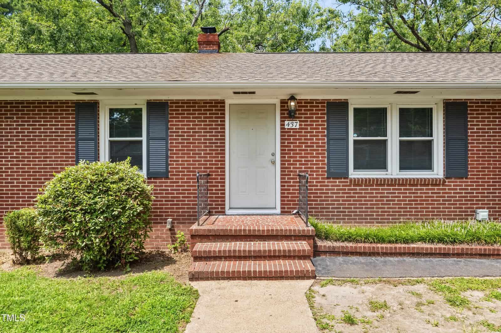A brick house with dark shutters, a white door, and small steps leading to the entrance. There is a bush to the left of the door and a grassy lawn in front.