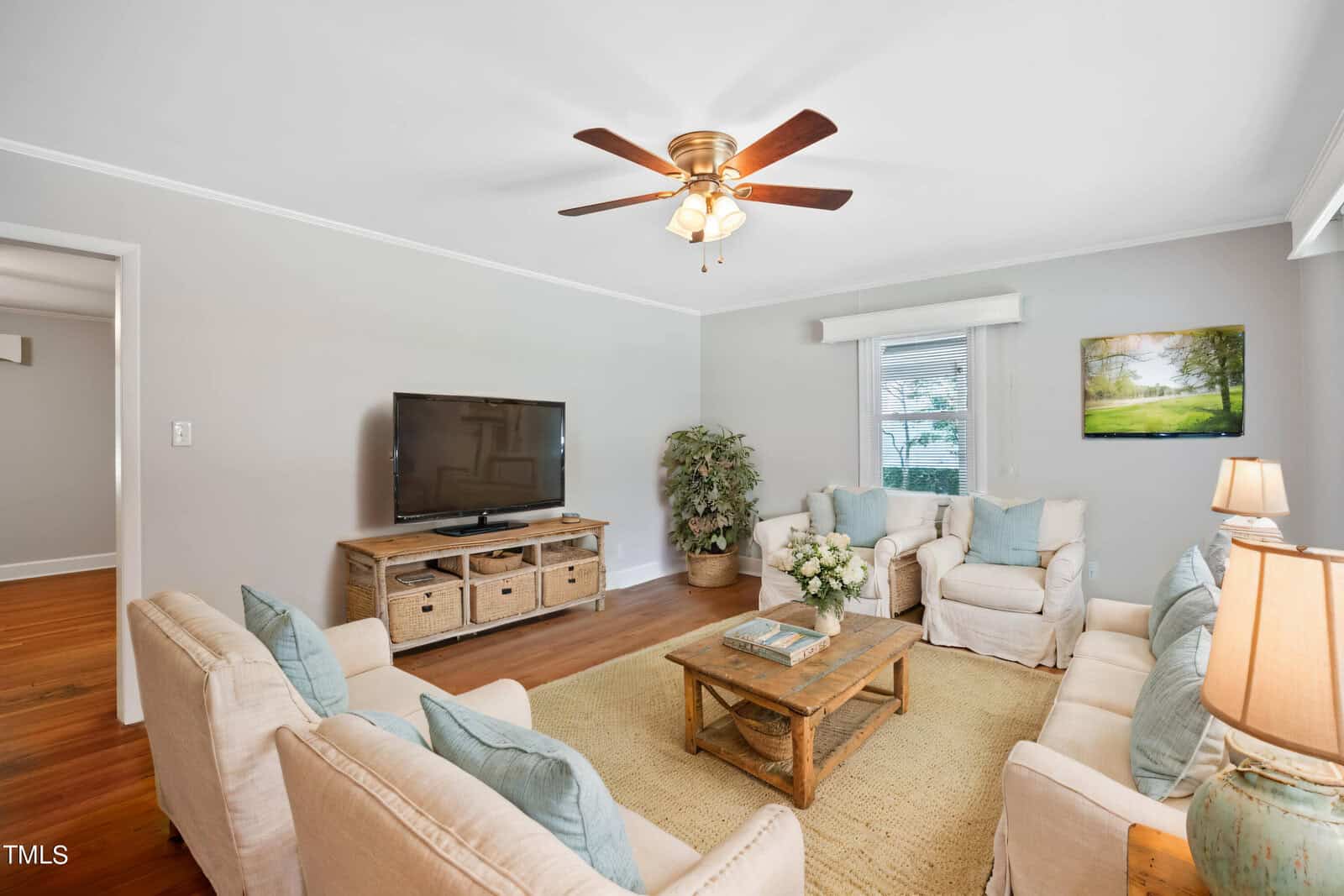 A cozy living room with light walls, beige sofas, a wooden coffee table, a TV on a stand with baskets, a potted plant, and a ceiling fan. Large windows bring in natural light, and soft decor accents the space.