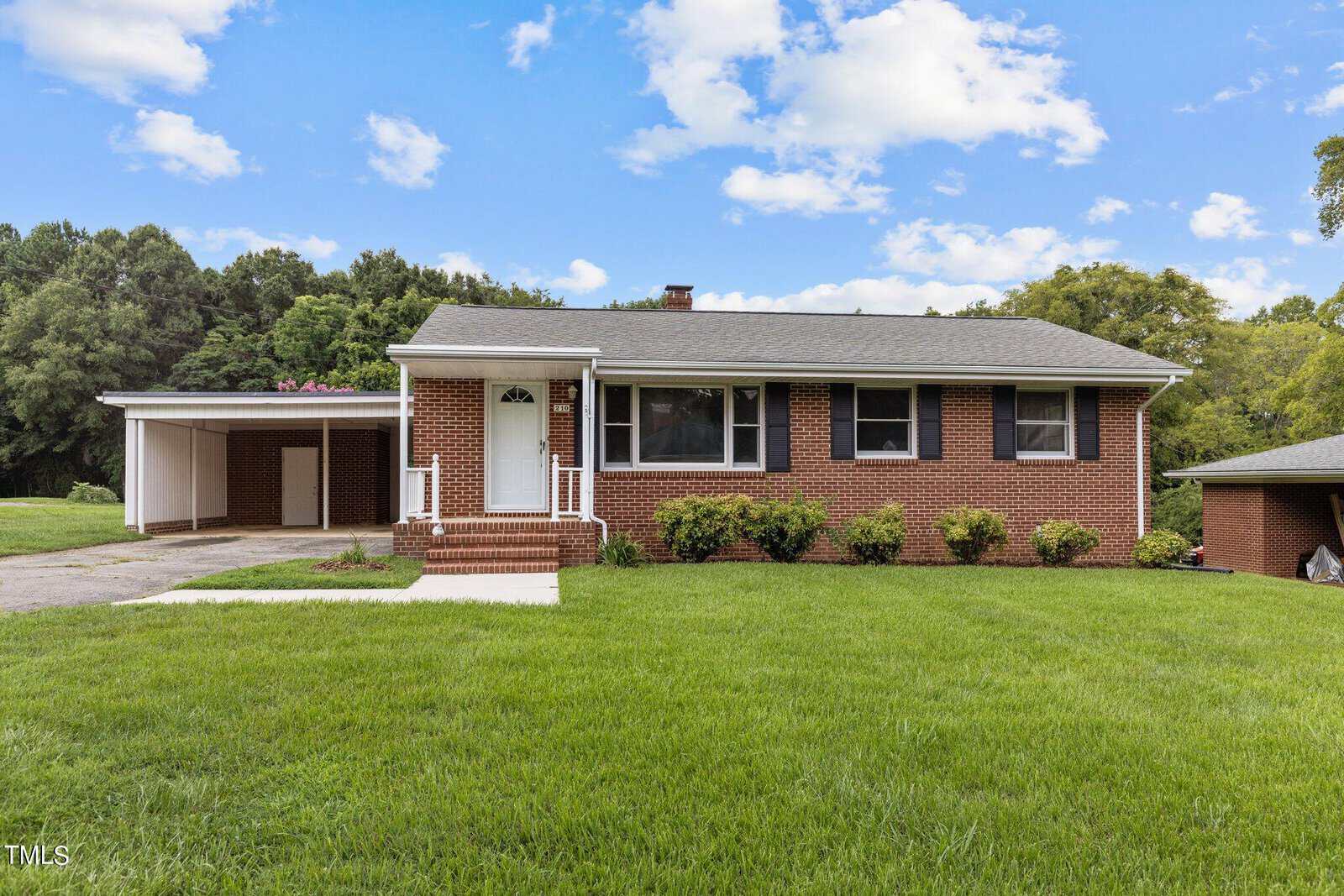 Nestled at 210 Grove Street, this charming brick house in Oxford features a large front yard, a convenient carport, and steps leading up to the welcoming front door. Surrounded by lush trees under a partly cloudy sky, it offers both tranquility and curb appeal.