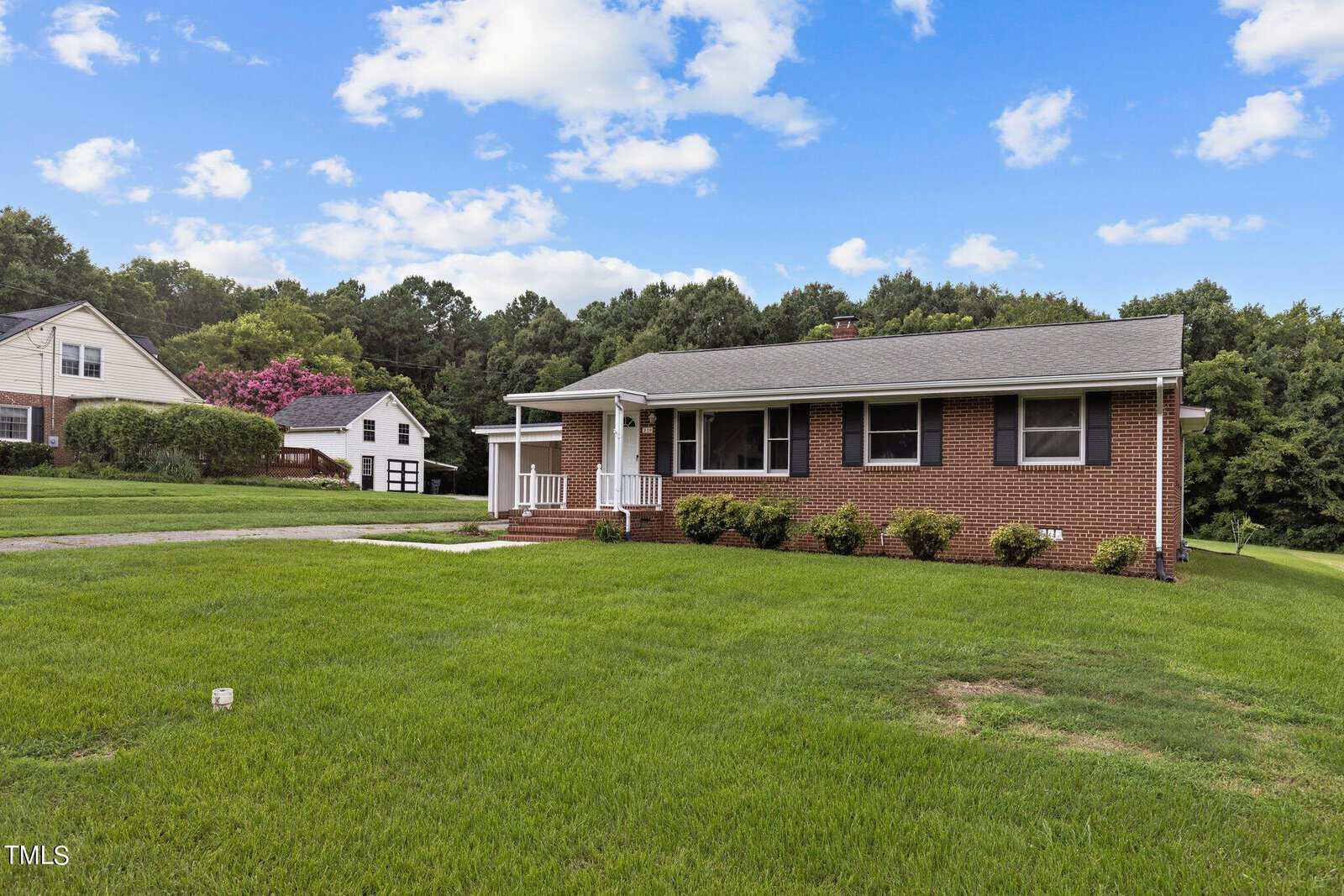 A one-story brick house with a gray roof is situated on a lush green lawn. The yard is bordered by trees, and a garage is visible in the background. A driveway leads to the front steps, and the sky is partly cloudy.