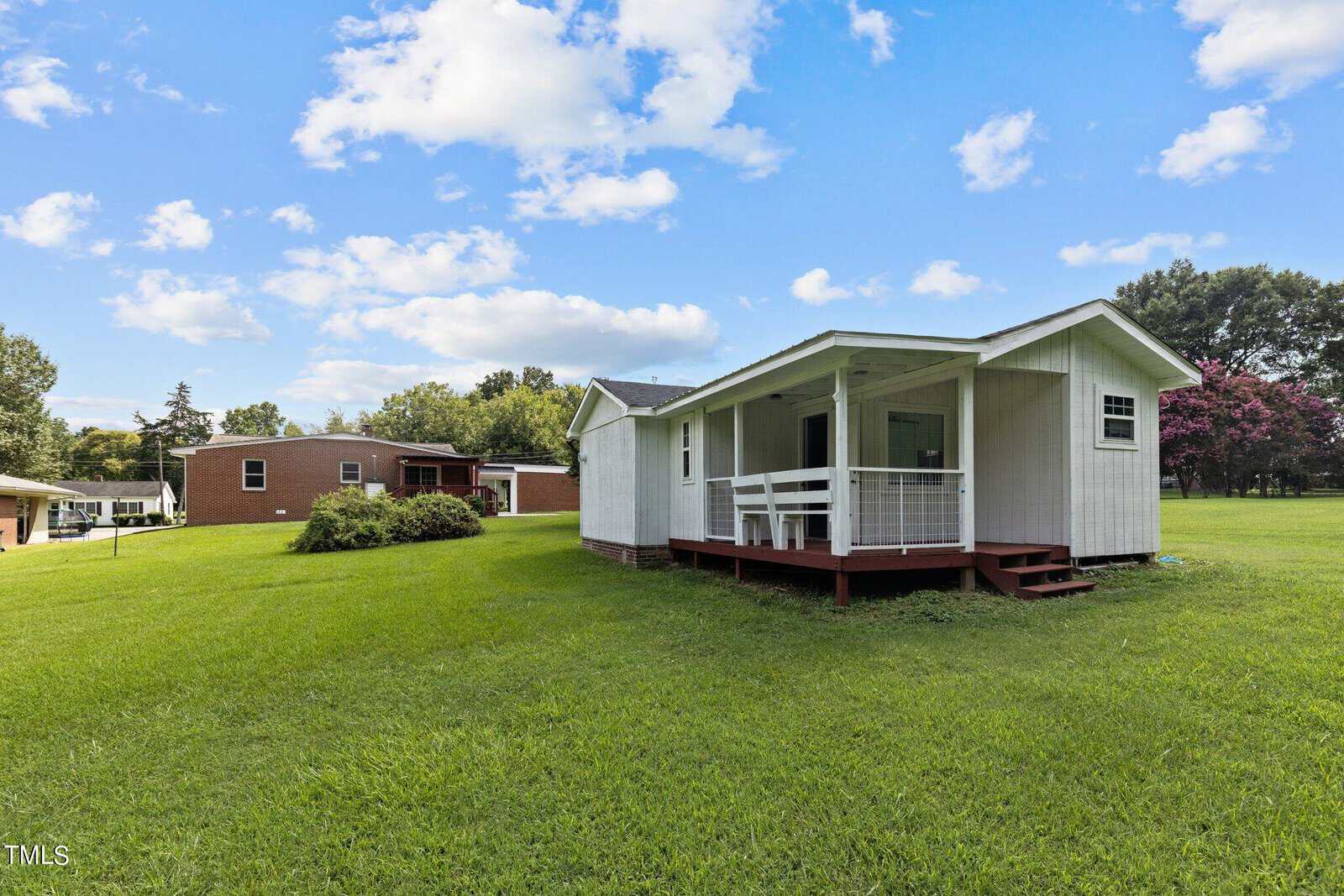 A small white cottage with a porch is set on a grassy lawn under a blue sky with scattered clouds. A larger red brick building is visible in the background, surrounded by trees and bushes.