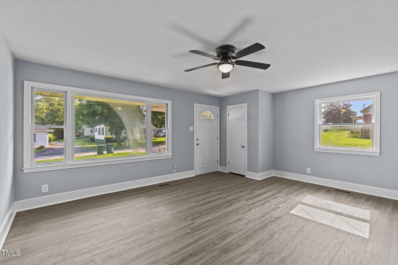 A bright living room with light gray walls and wood floor features a ceiling fan, large windows, and a white front door. Sunlight streams in, and the view outside shows greenery and neighboring houses.