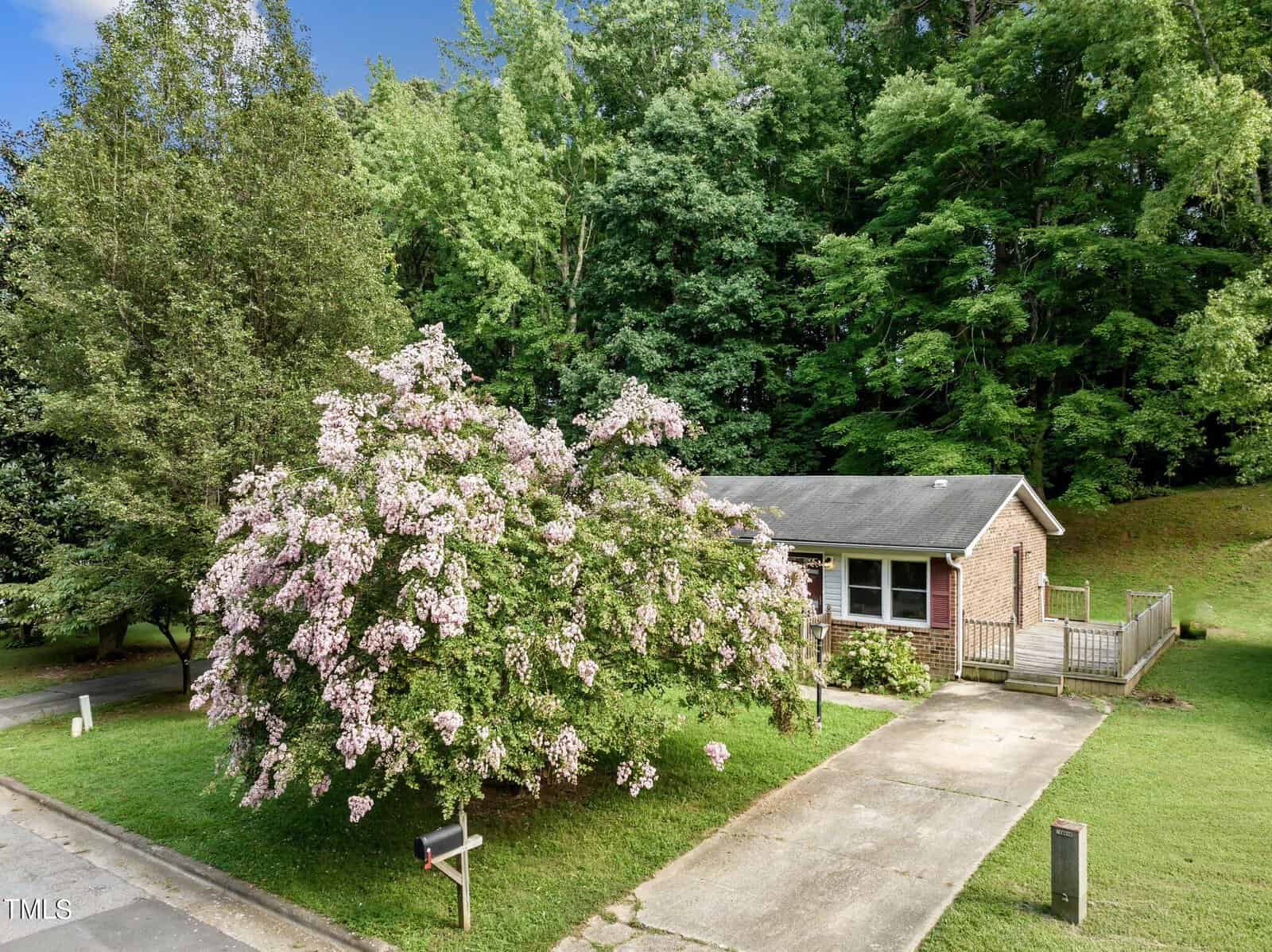 A small brick house with a sloped roof is surrounded by lush greenery and a blooming tree with pink flowers in the front yard. The driveway and mailbox are visible in the foreground under a clear blue sky.
