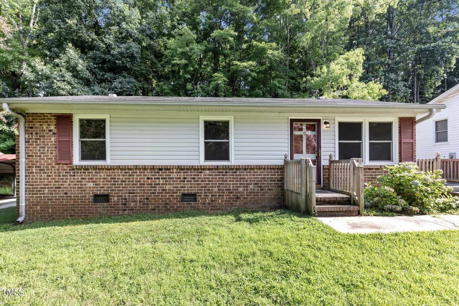 Located at 213 W Westbury Drive, Oxford, this charming single-story brick and white siding house boasts red shutters. It features a small wooden porch and ramp, surrounded by lush green grass and trees in the background.