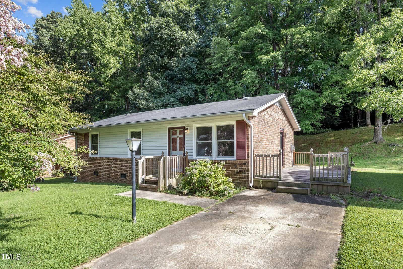A small, single-story brick house with a front porch and a wooden ramp. It is surrounded by lush green grass and tall trees. A driveway and a lamppost are in the foreground.