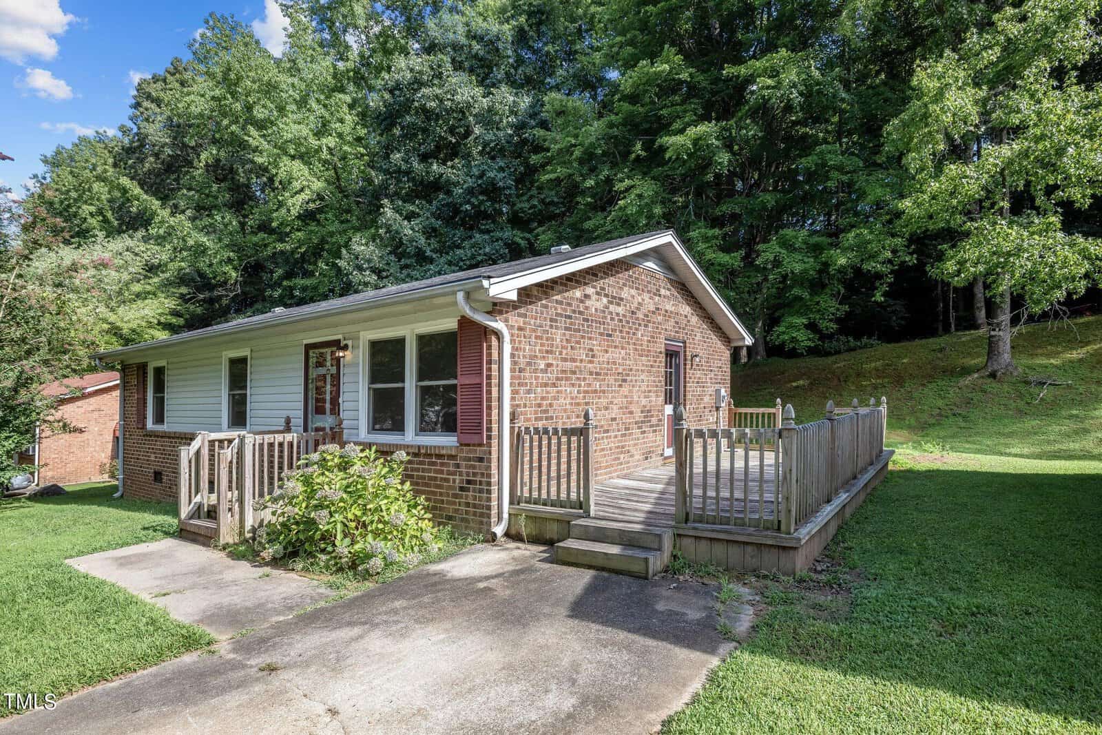 A single-story brick house with a wooden ramp leading to the front door. It's surrounded by a lush green yard and trees. The sky is clear and blue, indicating a sunny day.